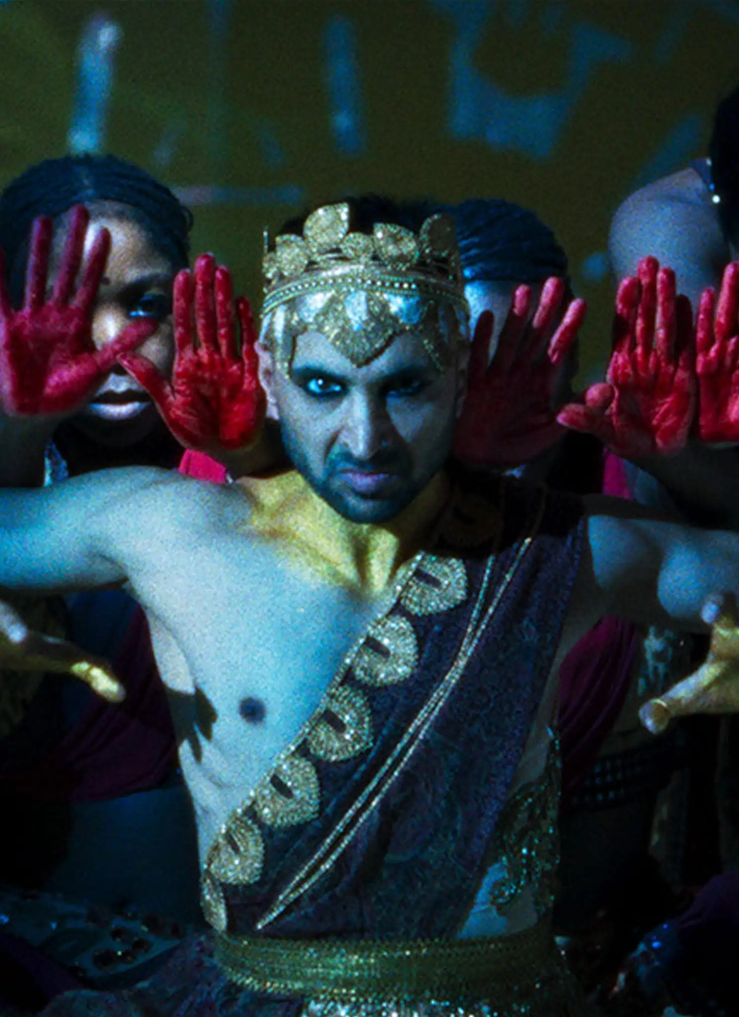 A performer in ornate golden attire and crown poses with outstretched hands, flanked by dancers holding up palms painted red, evoking a dramatic, synchronized stage performance with vivid lighting.