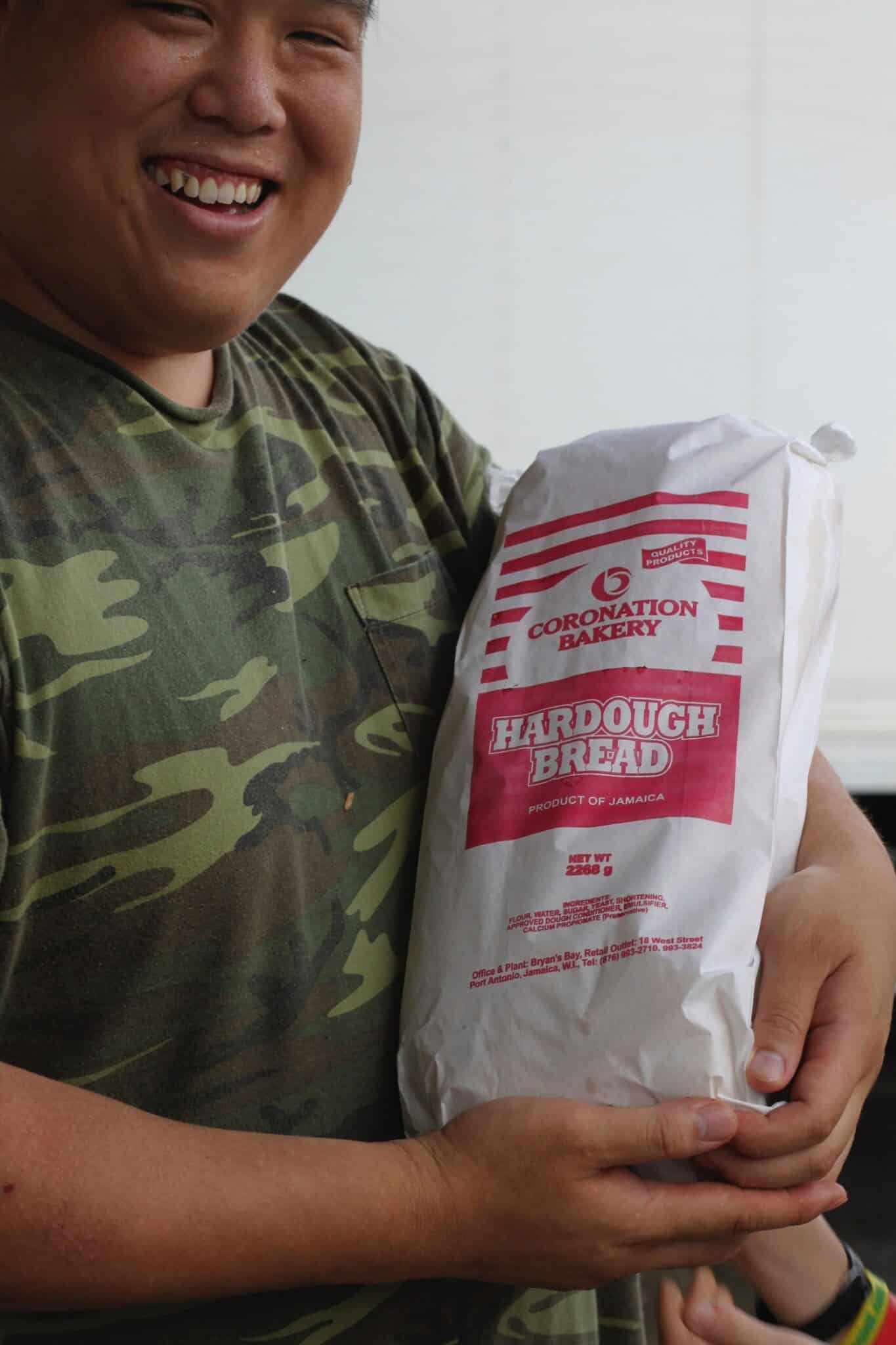 A person in a camouflage shirt smiles while holding a large loaf of Coronation Bakery Hardough Bread wrapped in white and red packaging.