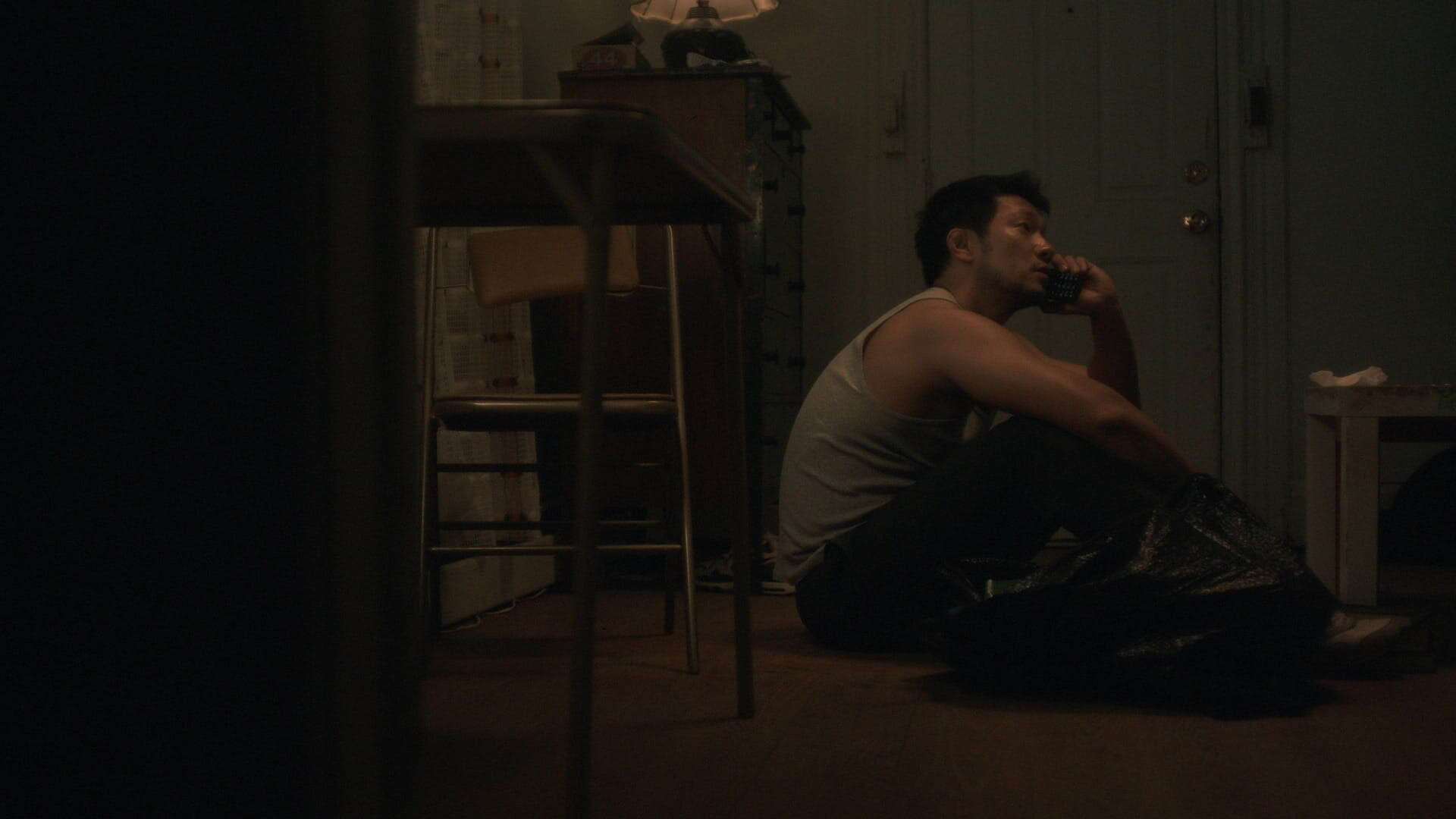 A man in a gray tank top sits on the floor of a dimly lit room, holding a phone to his ear. He is next to a trash bag, near a table and chair, with a dresser and closed door in the background.