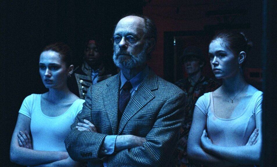 A serious-looking older man in a plaid suit stands with arms crossed between two young women in white leotards, also with arms crossed, under blue lighting, with other people blurred in the background.