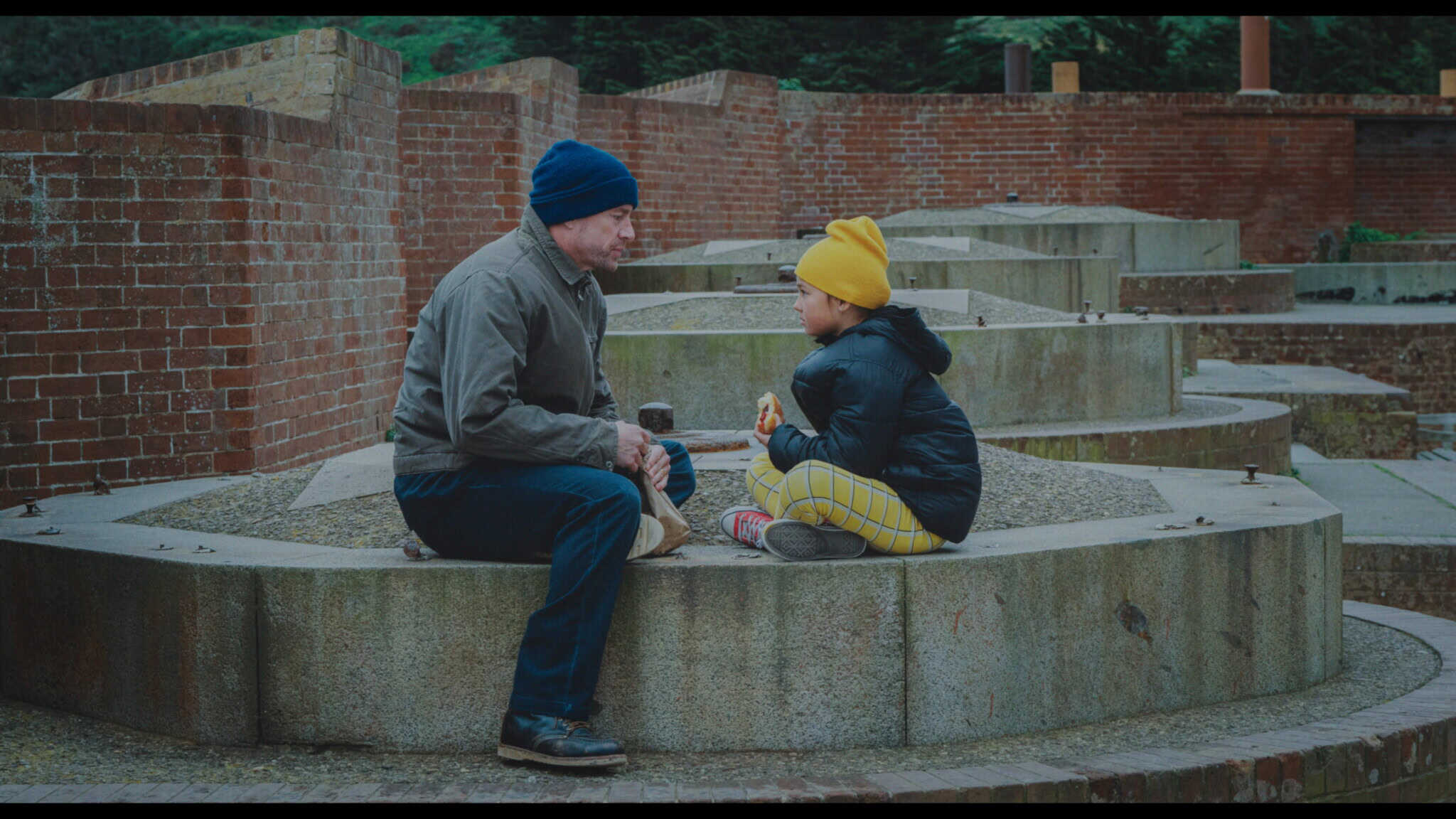 A man and a young girl, both wearing winter hats and jackets, sit facing each other on a curved concrete structure in an outdoor park. The girl holds a snack while they talk, surrounded by brick walls and greenery.