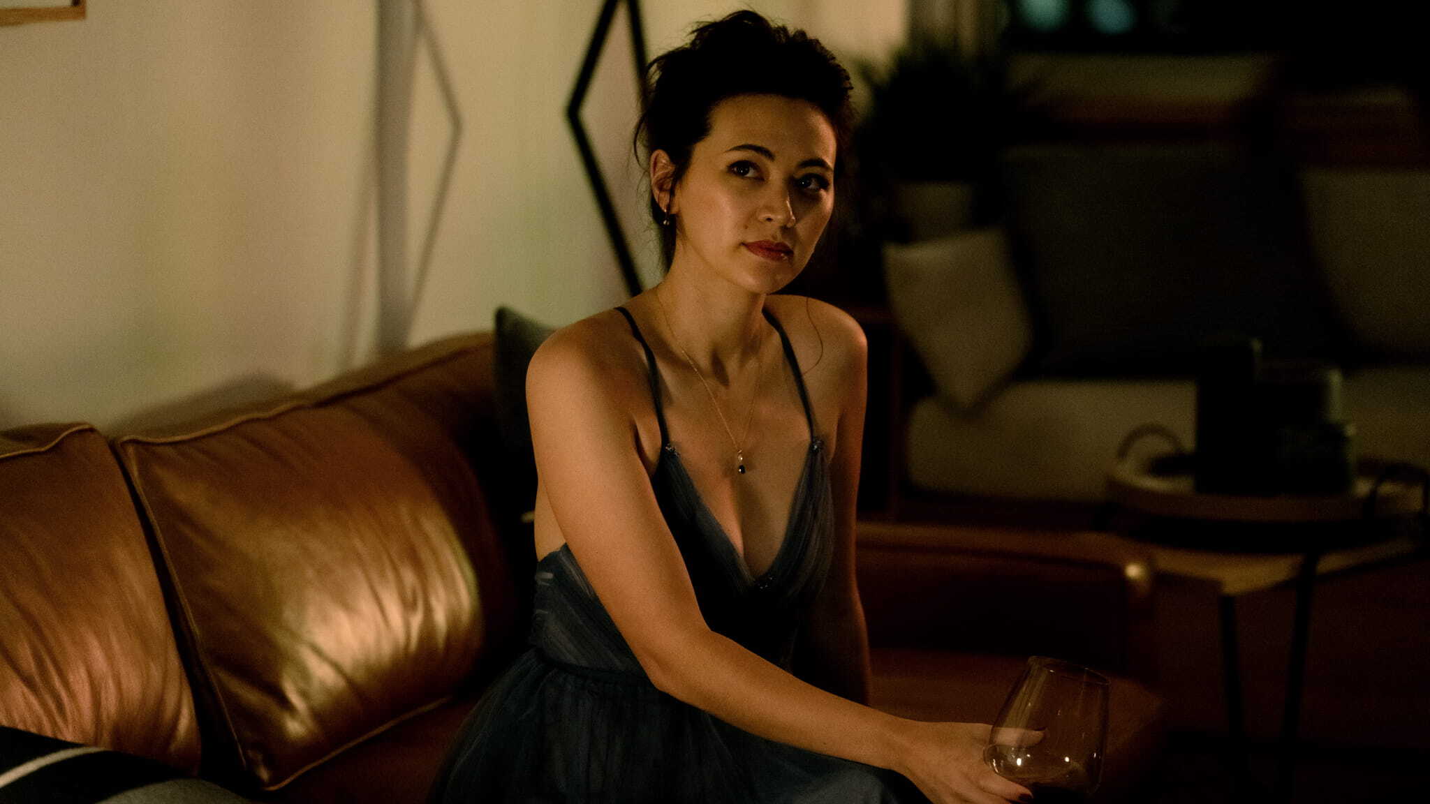 A woman in a blue dress sits on a brown leather couch in a dimly lit room, holding a wine glass and looking slightly to the side with a thoughtful expression.