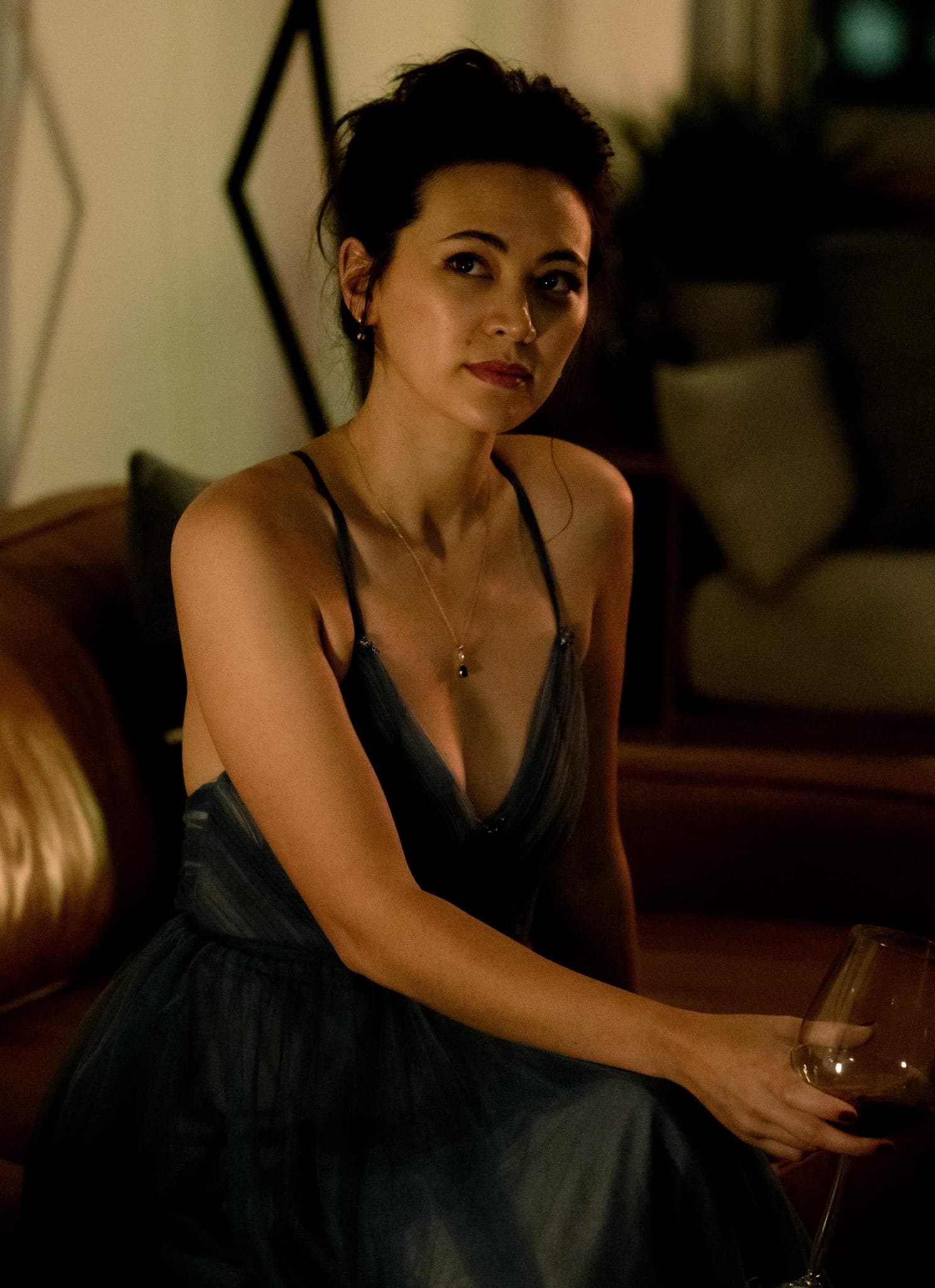 A woman in a blue dress sits on a brown leather couch in a dimly lit room, holding a wine glass and looking slightly to the side with a thoughtful expression.