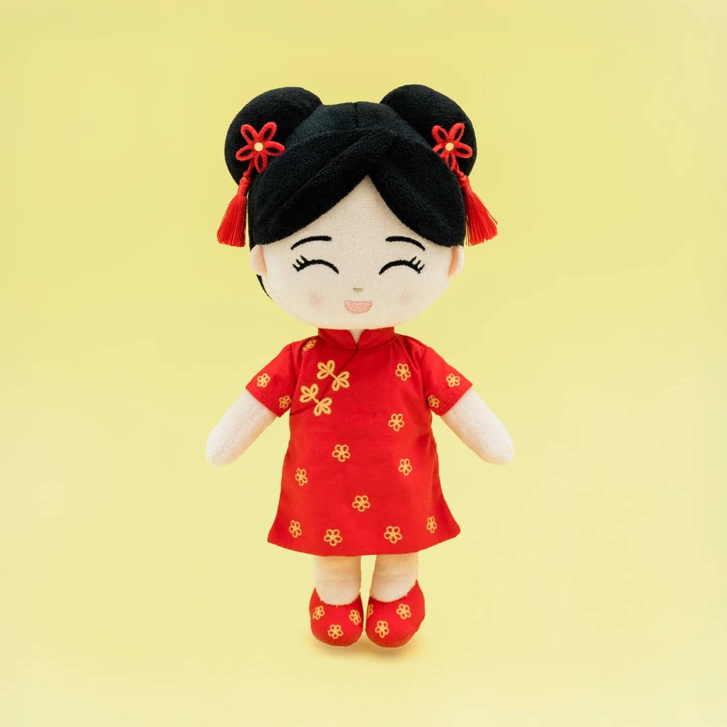 A plush doll with black hair styled in two buns, adorned with red accessories, celebrates Lunar New Year collections in a red dress with yellow floral patterns and matching shoes, smiling against a plain yellow background.