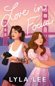 Illustrated cover of Love in Focus by Lyla Lee, featuring two young women standing in front of the Golden Gate Bridge. One holds a camera and wears a white top, the other wears a pink dress.