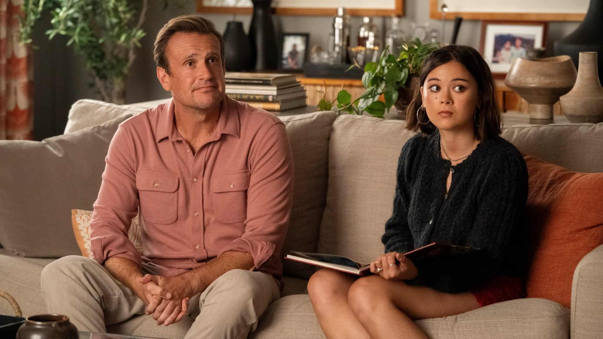 A man in a pink button-up shirt and a young woman holding an open book sit side by side on a beige couch, both looking slightly off-camera with serious expressions in a cozy living room setting.