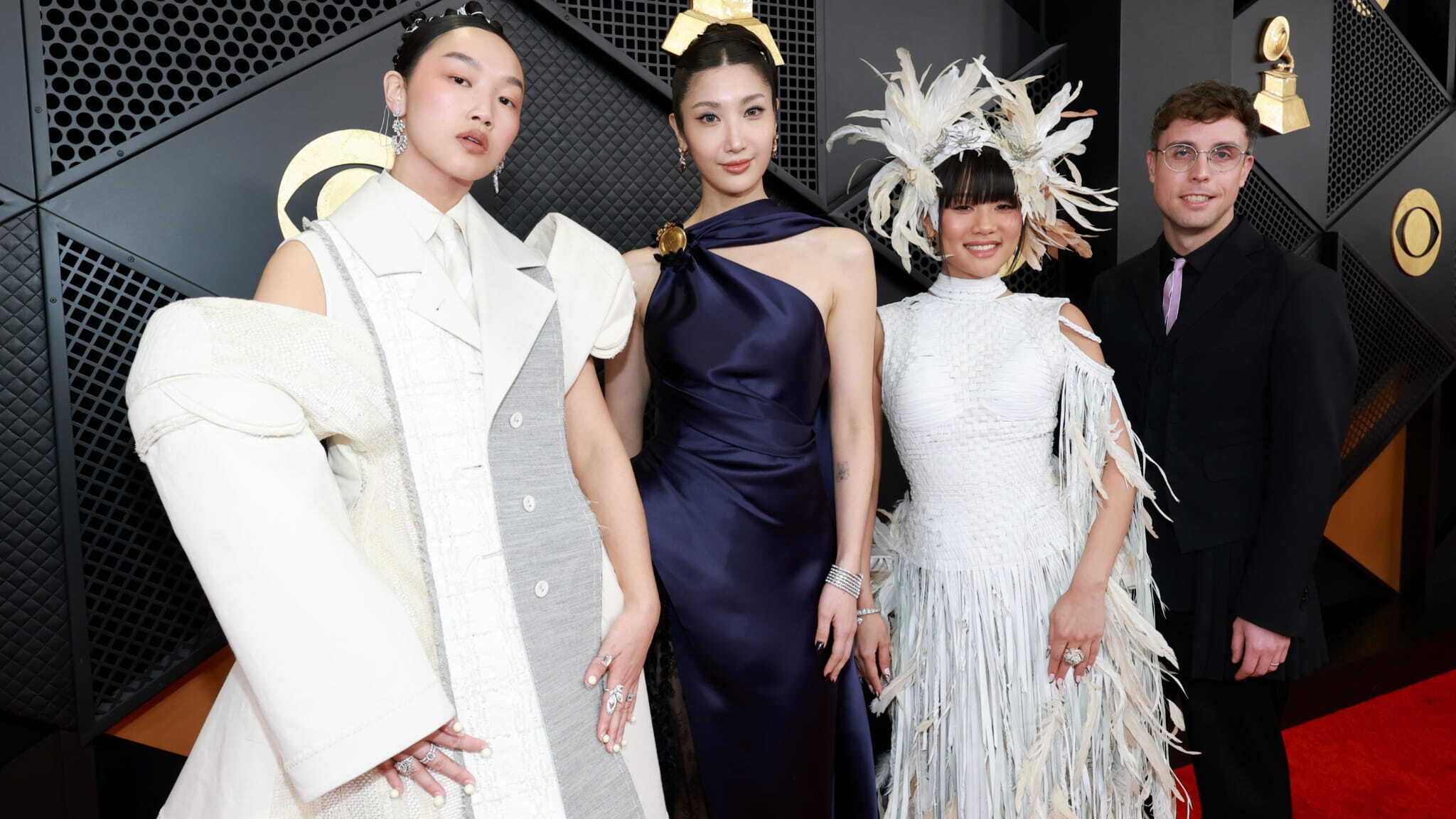 Four people pose on a red carpet in stylish, avant-garde outfits. One wears a layered white suit, another a satin navy gown, the third a white feathered dress with a headpiece, and the fourth a black suit with glasses.