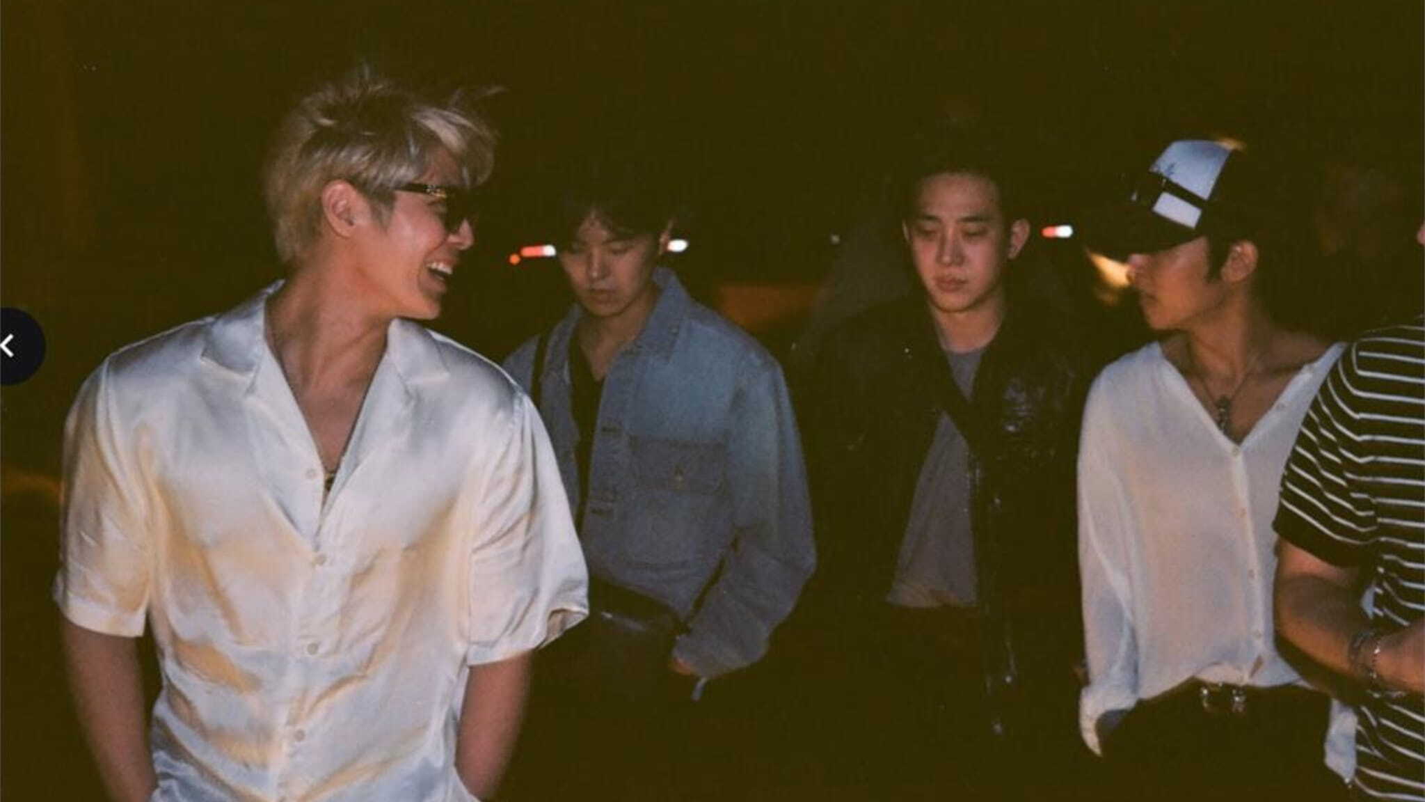 Four young men walk together at night, talking and smiling under dim lighting. Two wear white shirts, one wears a light denim jacket, and one sports a dark jacket—reminiscent of The Rose's effortless style.