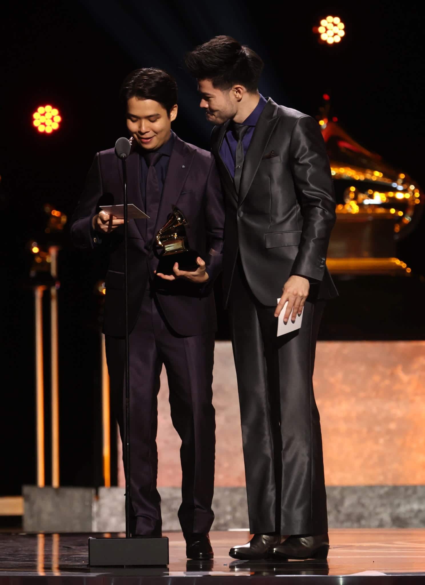 Two men in suits stand on stage at the Grammys, one holding a Grammy trophy and reading from a phone as the other looks on. Bright lights and a large Grammy statue frame the moment, celebrating ARKAI’s achievement.