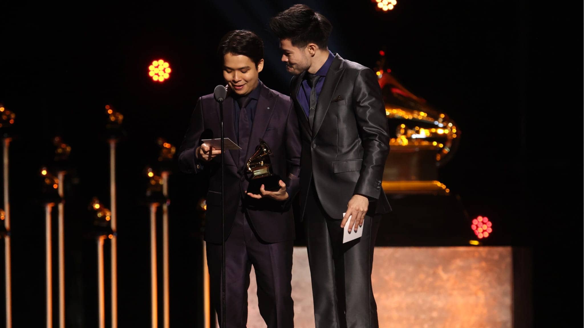 Two men in suits stand on stage at the Grammys, one holding a Grammy trophy and reading from a phone as the other looks on. Bright lights and a large Grammy statue frame the moment, celebrating ARKAI’s achievement.