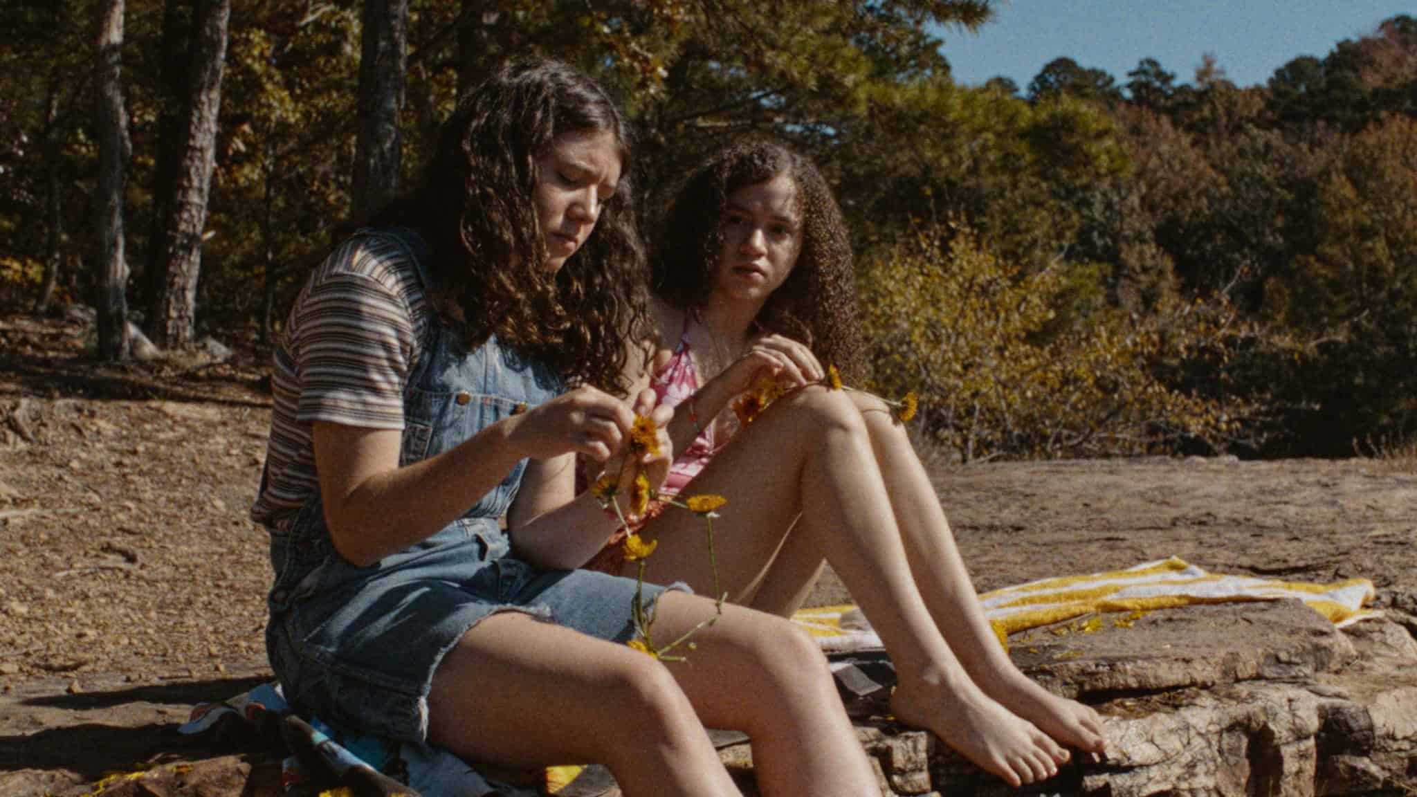 Two girls sit on a rock outdoors near trees. One girl in overalls examines a branch, while the other, in a pink top, sits beside her with her legs stretched out on a towel. Both appear thoughtful and focused.