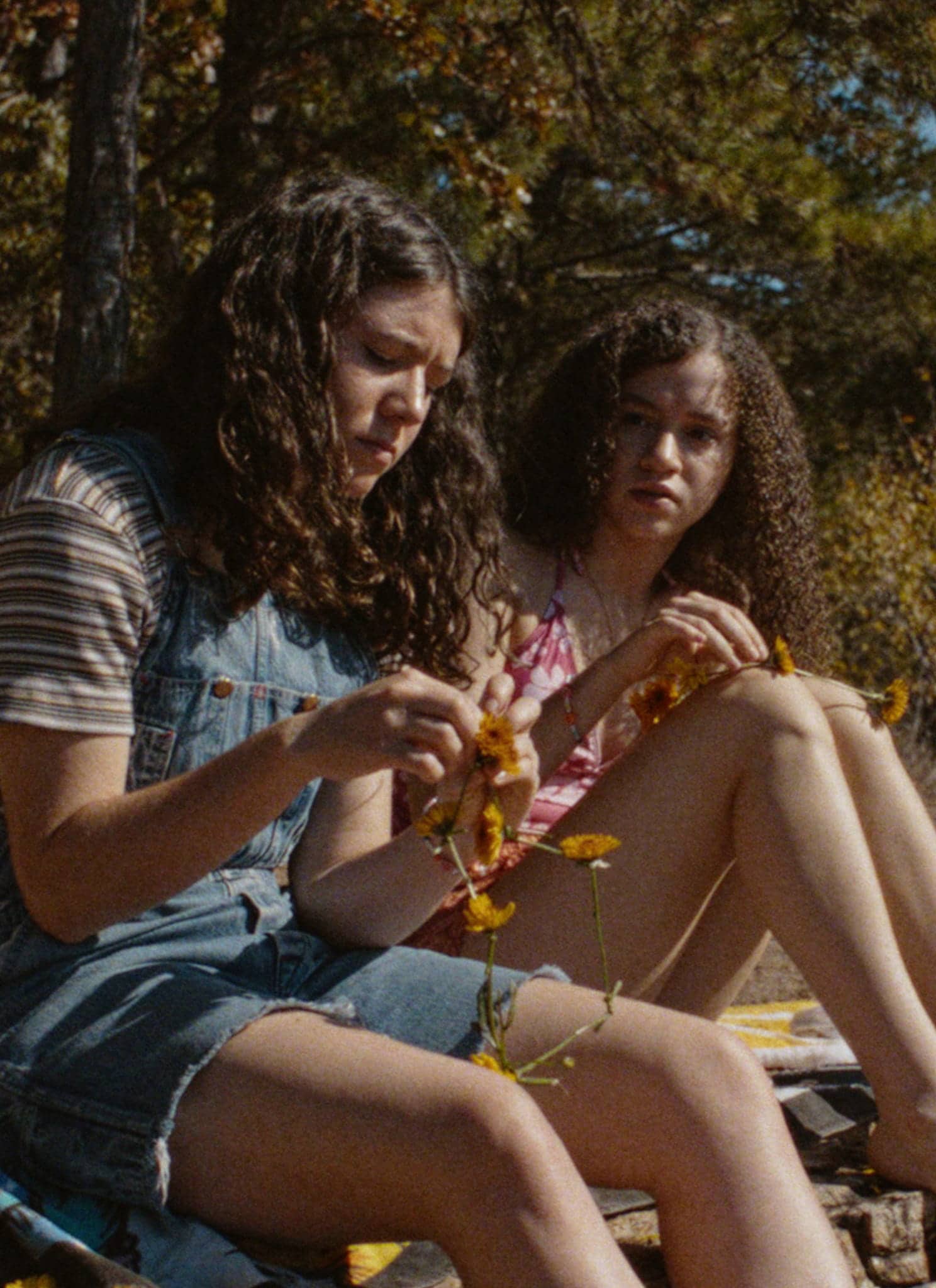 Two girls sit on a rock outdoors near trees. One girl in overalls examines a branch, while the other, in a pink top, sits beside her with her legs stretched out on a towel. Both appear thoughtful and focused.