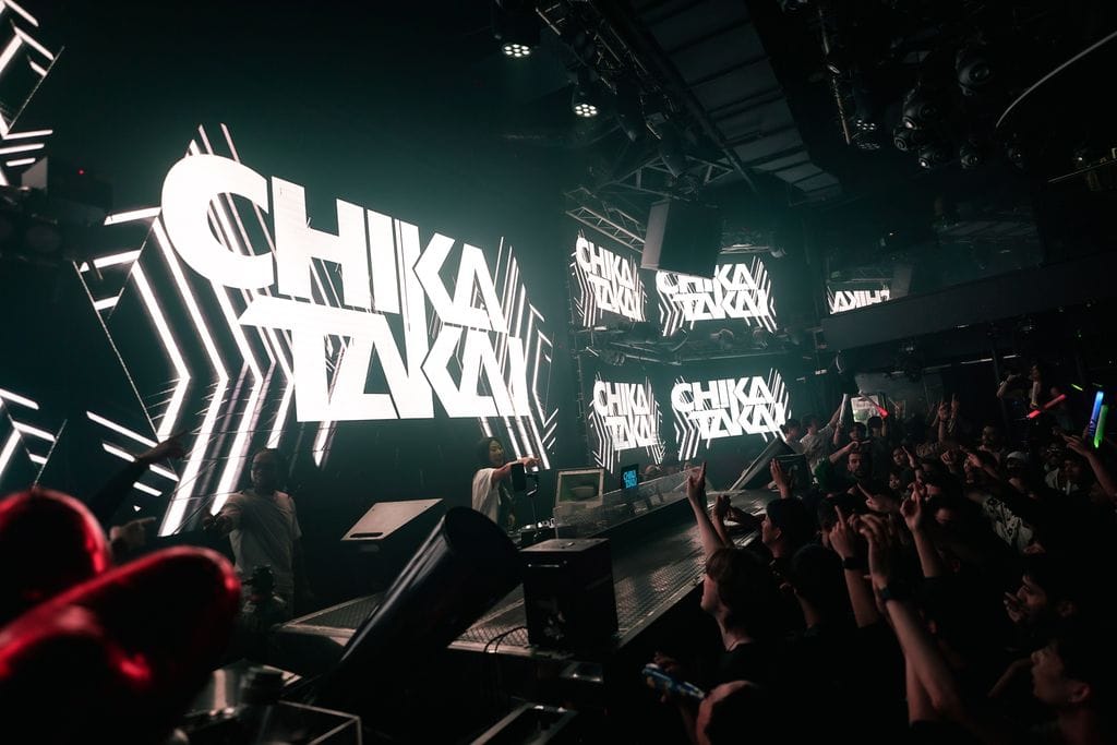 A crowded nightclub with a large LED screen displaying CHIKA TAKAI in bold white letters. People in the audience raise their hands and colorful lights illuminate the energetic scene.