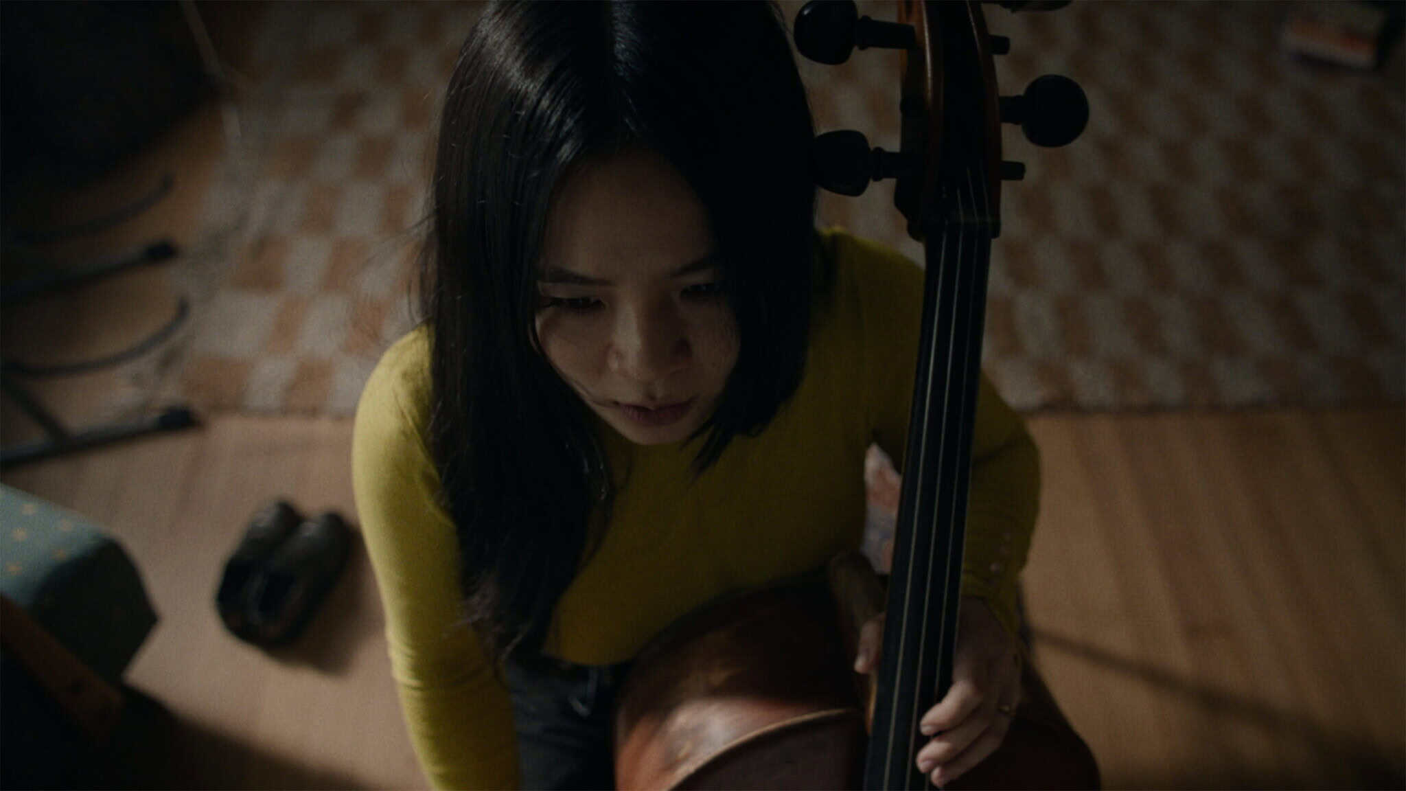 A woman with long dark hair, wearing a yellow top, sits and holds a cello, looking downward in a dimly lit room with a patterned rug and wooden floor.