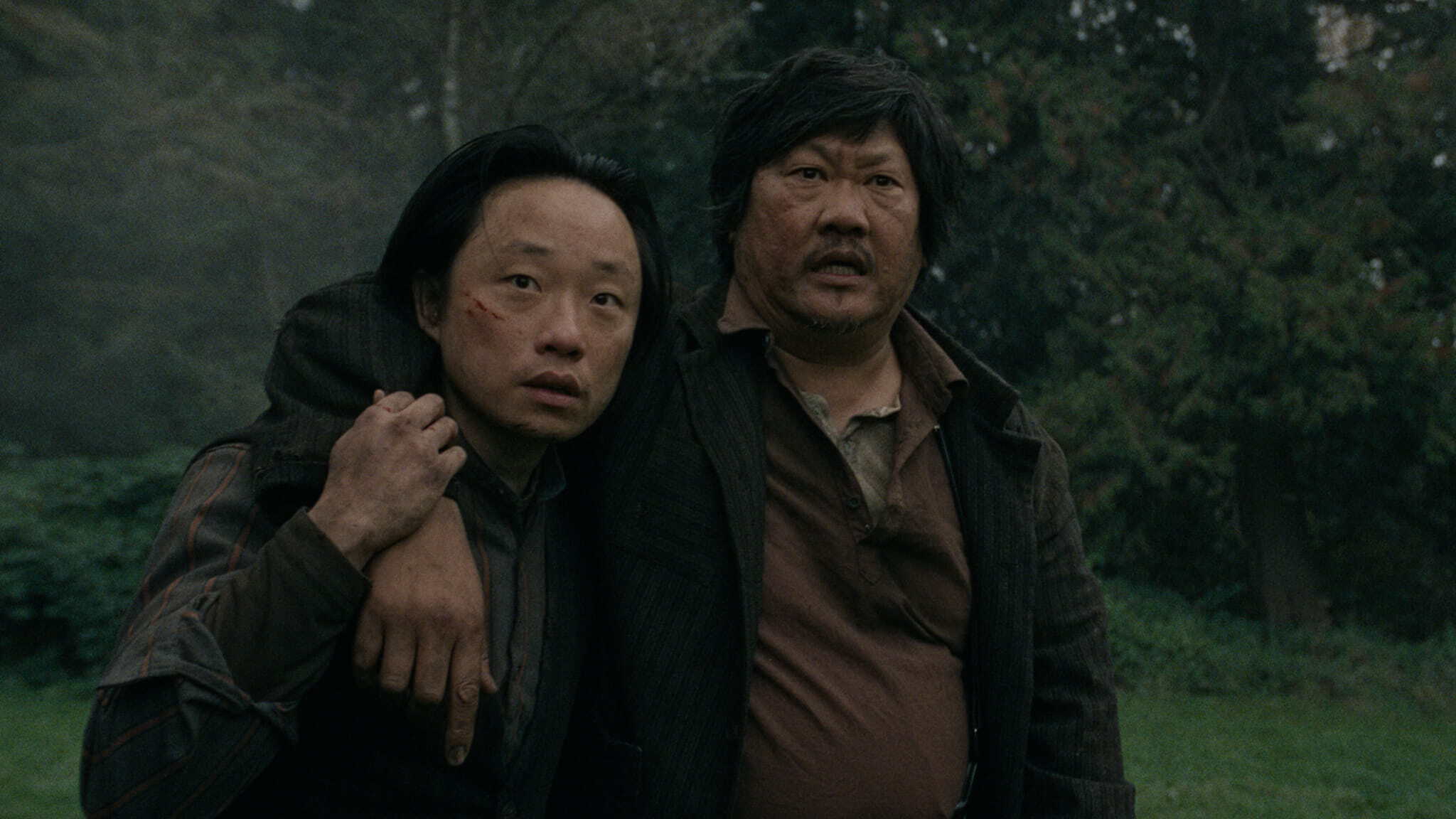 Two men stand outdoors in a wooded area. One man is injured with a scrape on his face, leaning heavily with his arm around the other mans shoulders for support. Both look concerned and are wearing dark, rugged clothing.