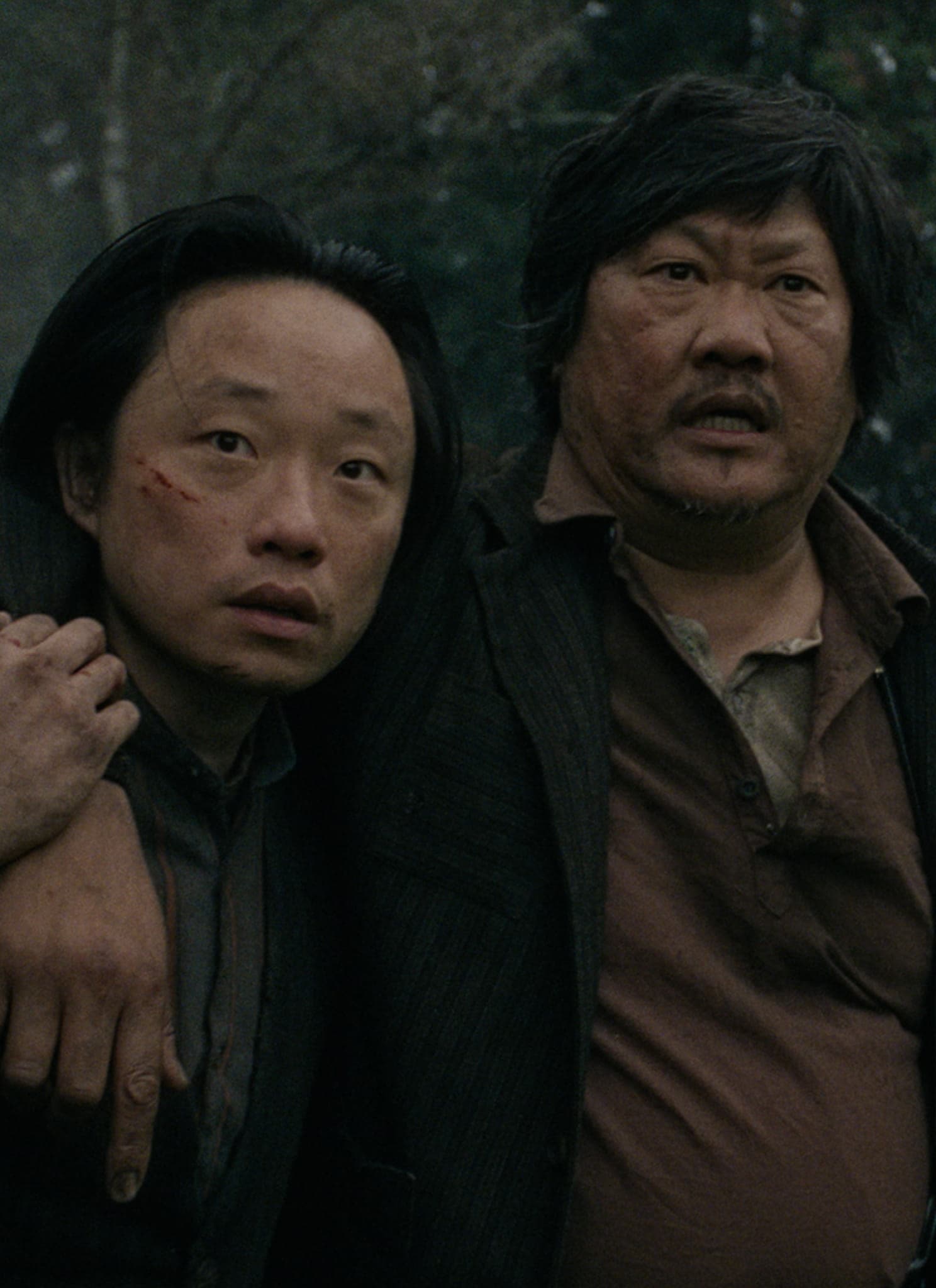 Two men stand outdoors in a wooded area. One man is injured with a scrape on his face, leaning heavily with his arm around the other mans shoulders for support. Both look concerned and are wearing dark, rugged clothing.