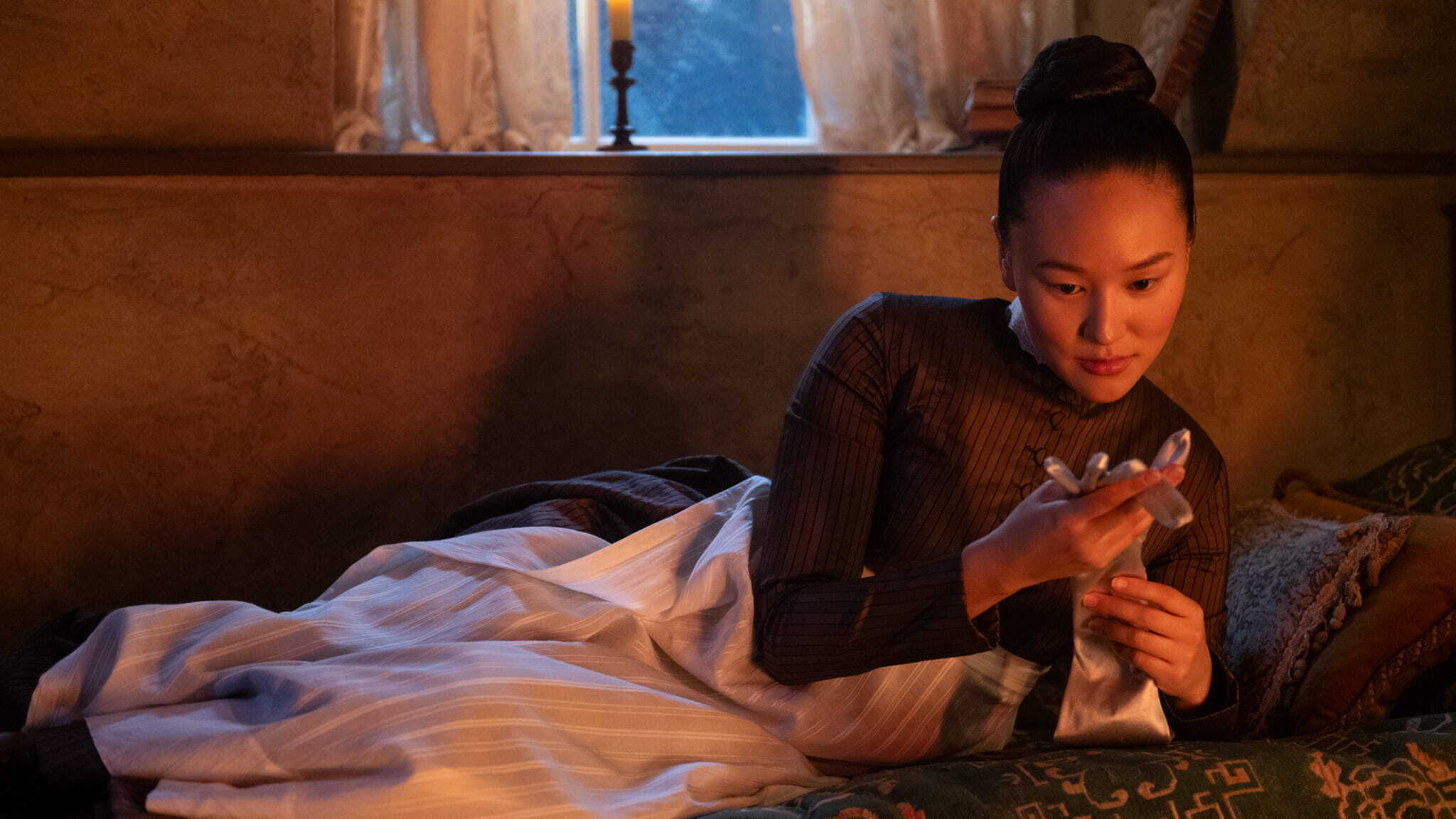 A woman in period clothing reclines on a bed, thoughtfully looking at a pair of gloves in her hands. A candle burns on the windowsill behind her, casting a warm light in the dimly lit room.