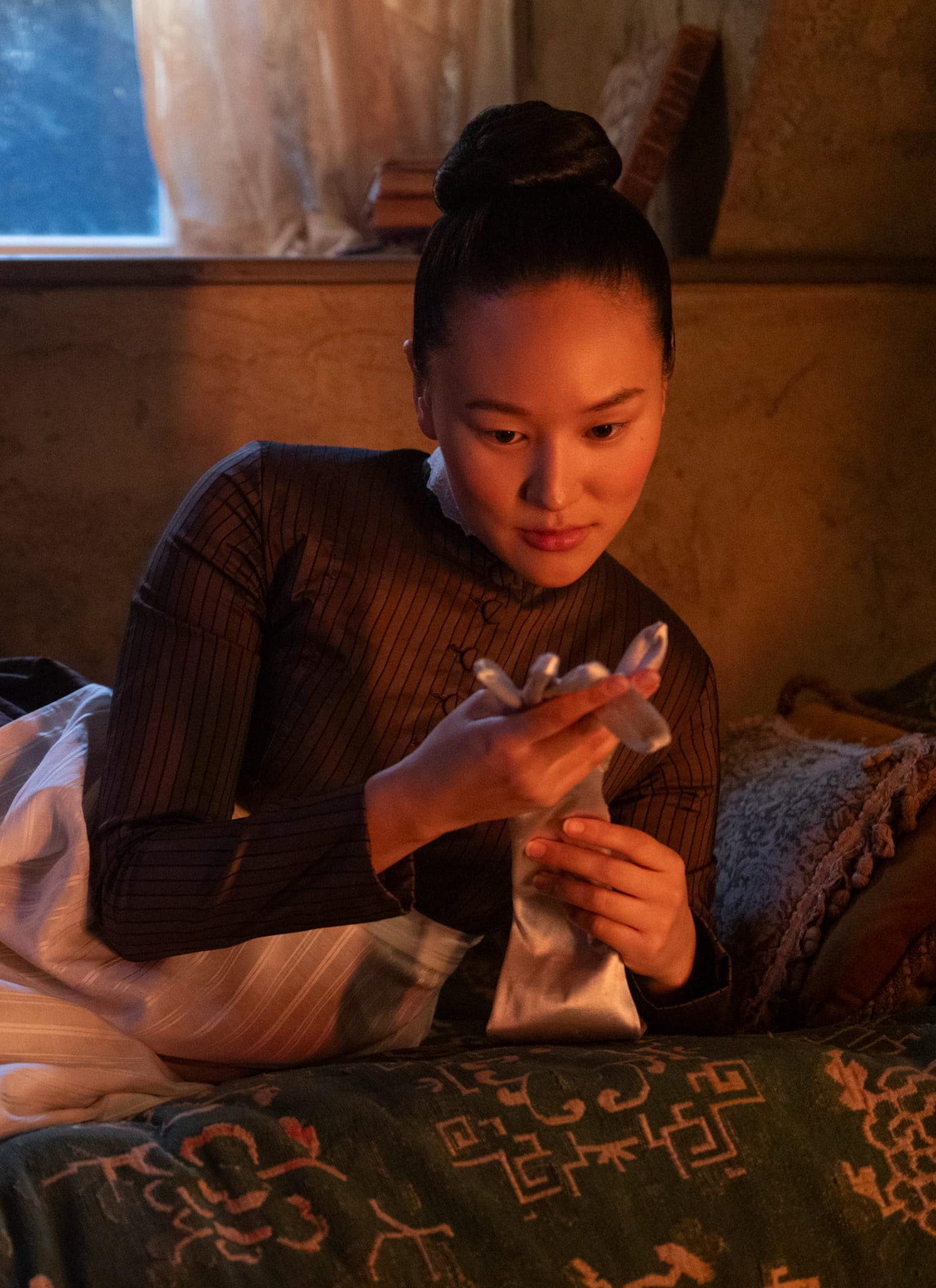 A woman in period clothing reclines on a bed, thoughtfully looking at a pair of gloves in her hands. A candle burns on the windowsill behind her, casting a warm light in the dimly lit room.