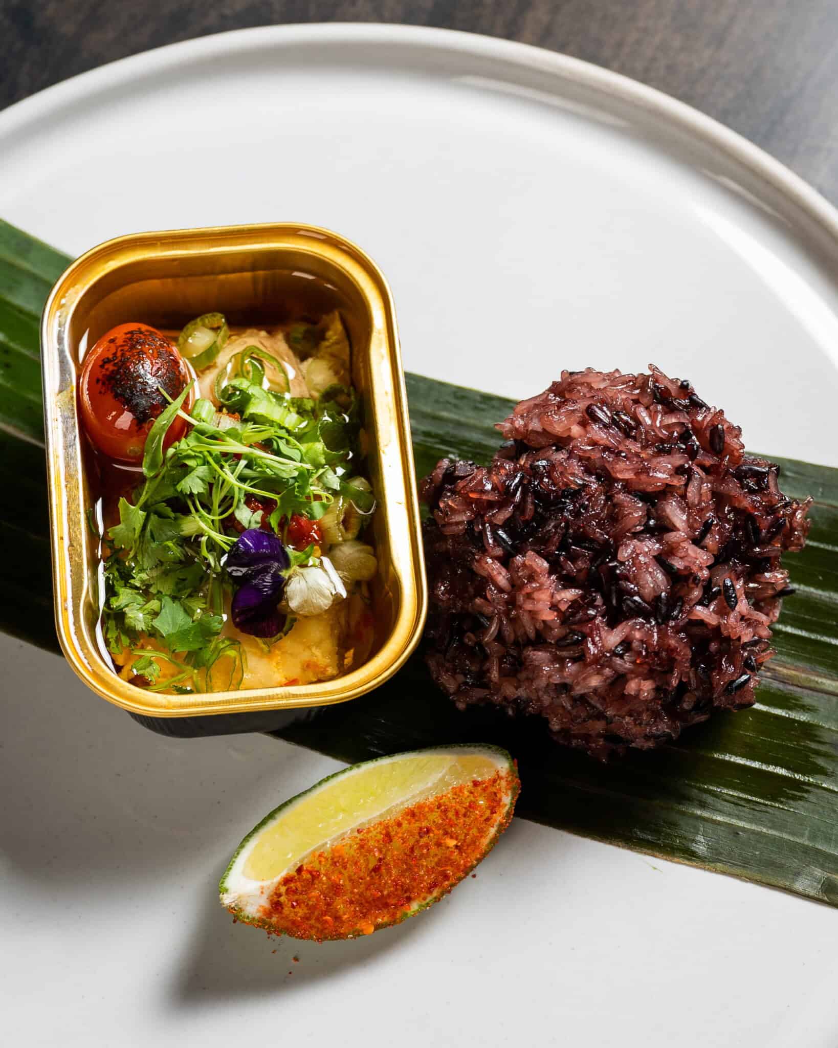 A plated dish inspired by Asian childhood snacks, featuring a tin container of colorful vegetables and herbs, a mound of black rice on a banana leaf, and a chili-dusted lime wedge on a white plate.