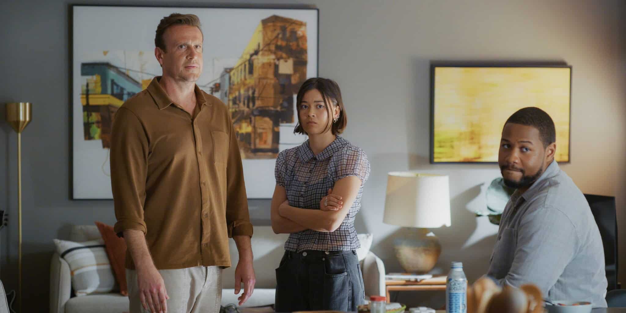 Three people are in a modern living room: a man in a brown shirt stands on the left, a woman with arms crossed stands in the middle, and a man in a blue shirt sits at a table on the right. They appear to be focused and serious.