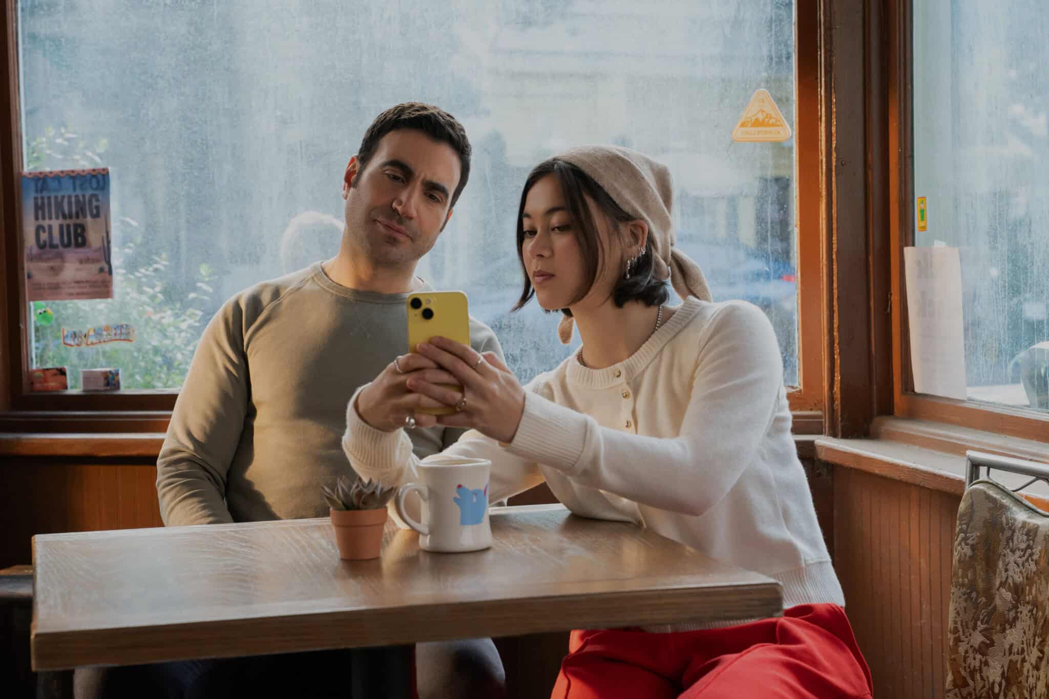 A woman and man sit at a café table by a window. The woman shows the man something on her yellow smartphone. Both appear focused. There’s a coffee mug and a small plant on the table. Posters are visible on the window.