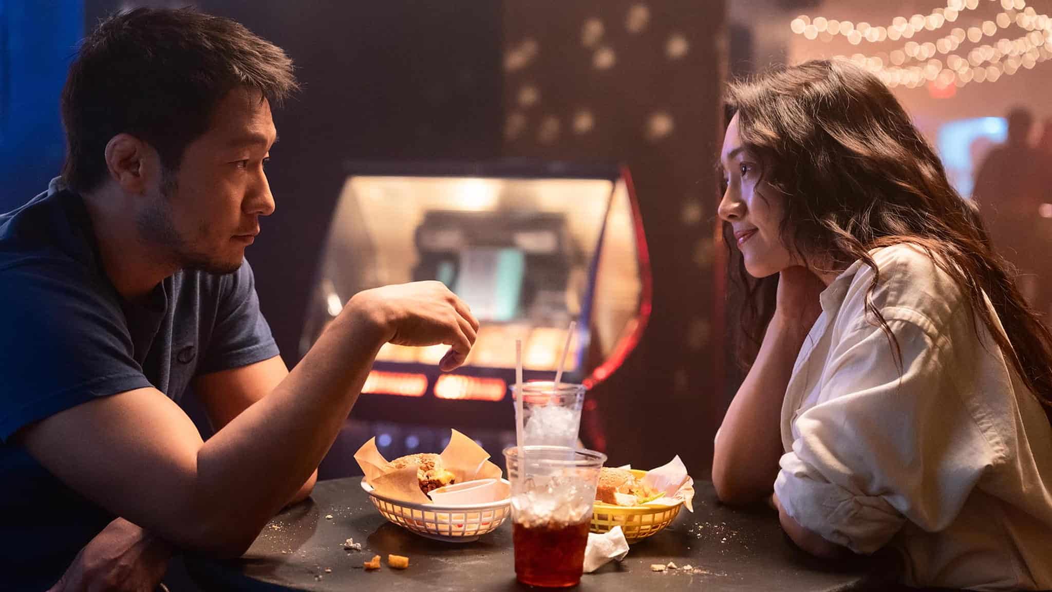 A man and a woman sit across from each other at a small table, sharing food and drinks in a dimly lit setting with string lights in the background. They look engaged in conversation.