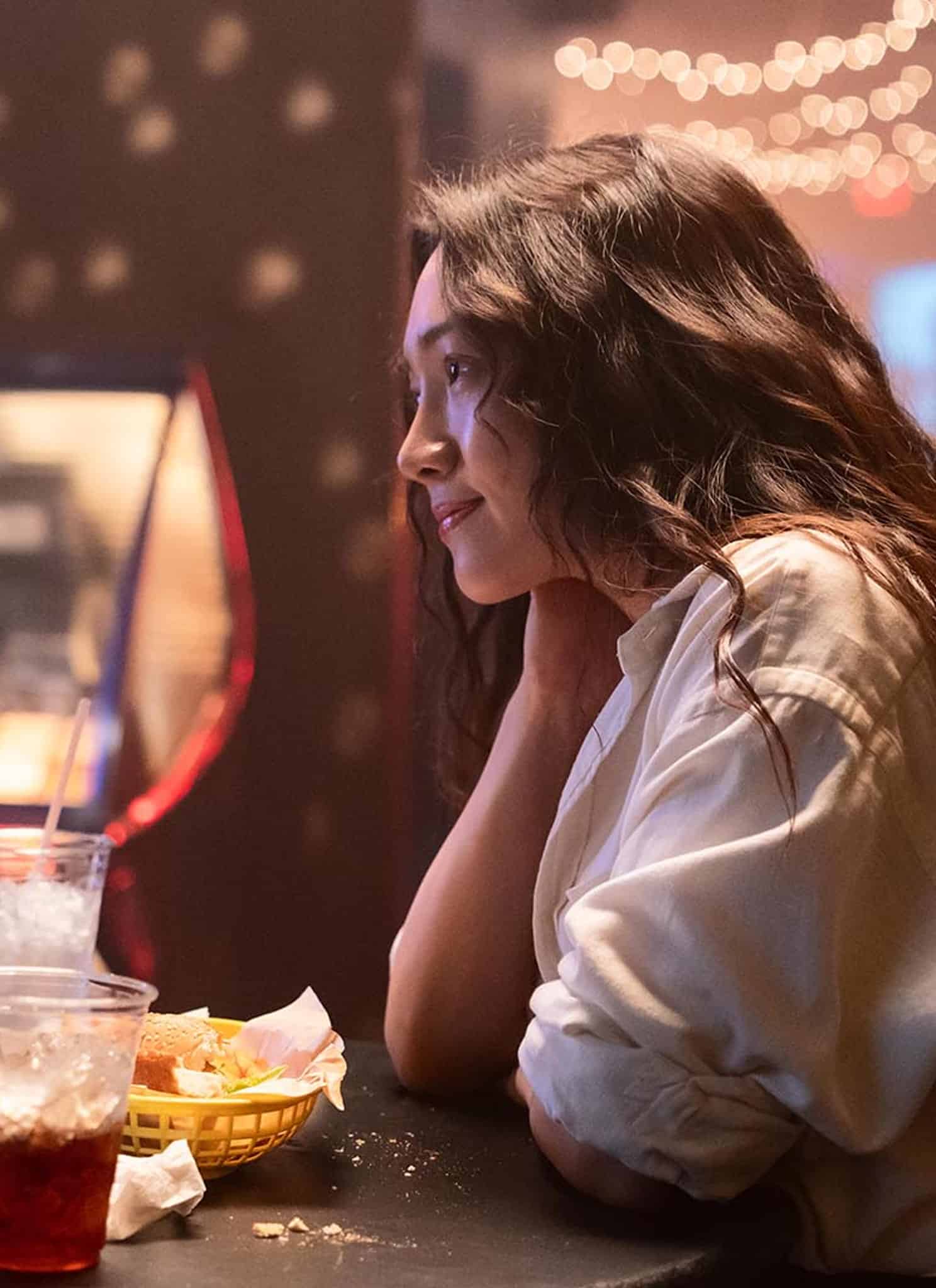 A man and a woman sit across from each other at a small table, sharing food and drinks in a dimly lit setting with string lights in the background. They look engaged in conversation.