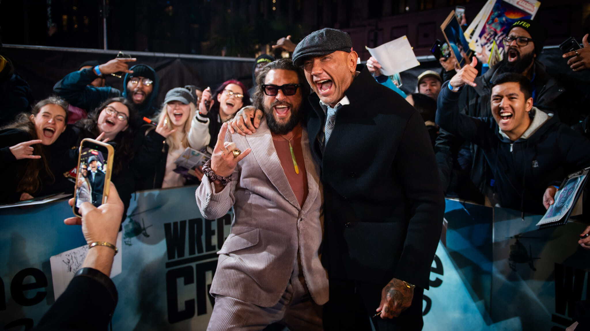 Two men pose excitedly on a blue carpet, smiling and hugging, surrounded by cheering fans holding phones and posters at an event. The atmosphere is lively and celebratory.