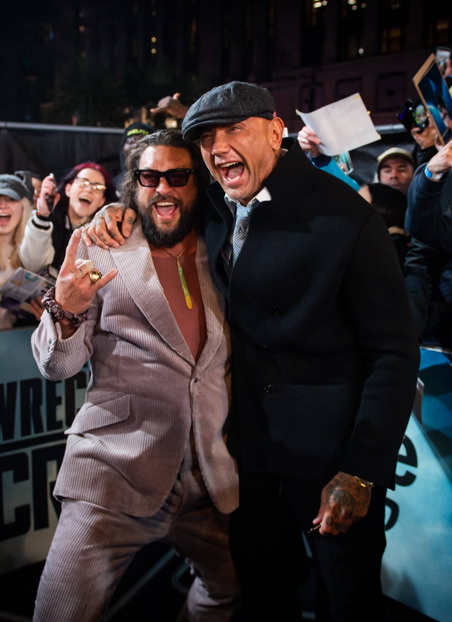 Two men pose excitedly on a blue carpet, smiling and hugging, surrounded by cheering fans holding phones and posters at an event. The atmosphere is lively and celebratory.
