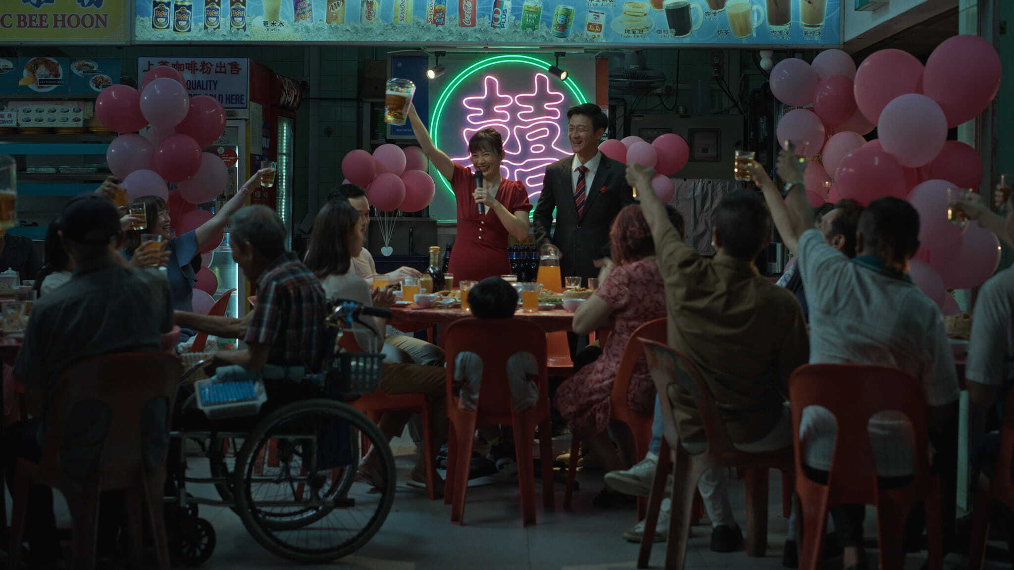 A group of people sit around tables decorated with pink balloons, raising glasses in a toast. Two people stand at the front, smiling, under a bright neon sign with double happiness symbols. The scene appears festive and celebratory.