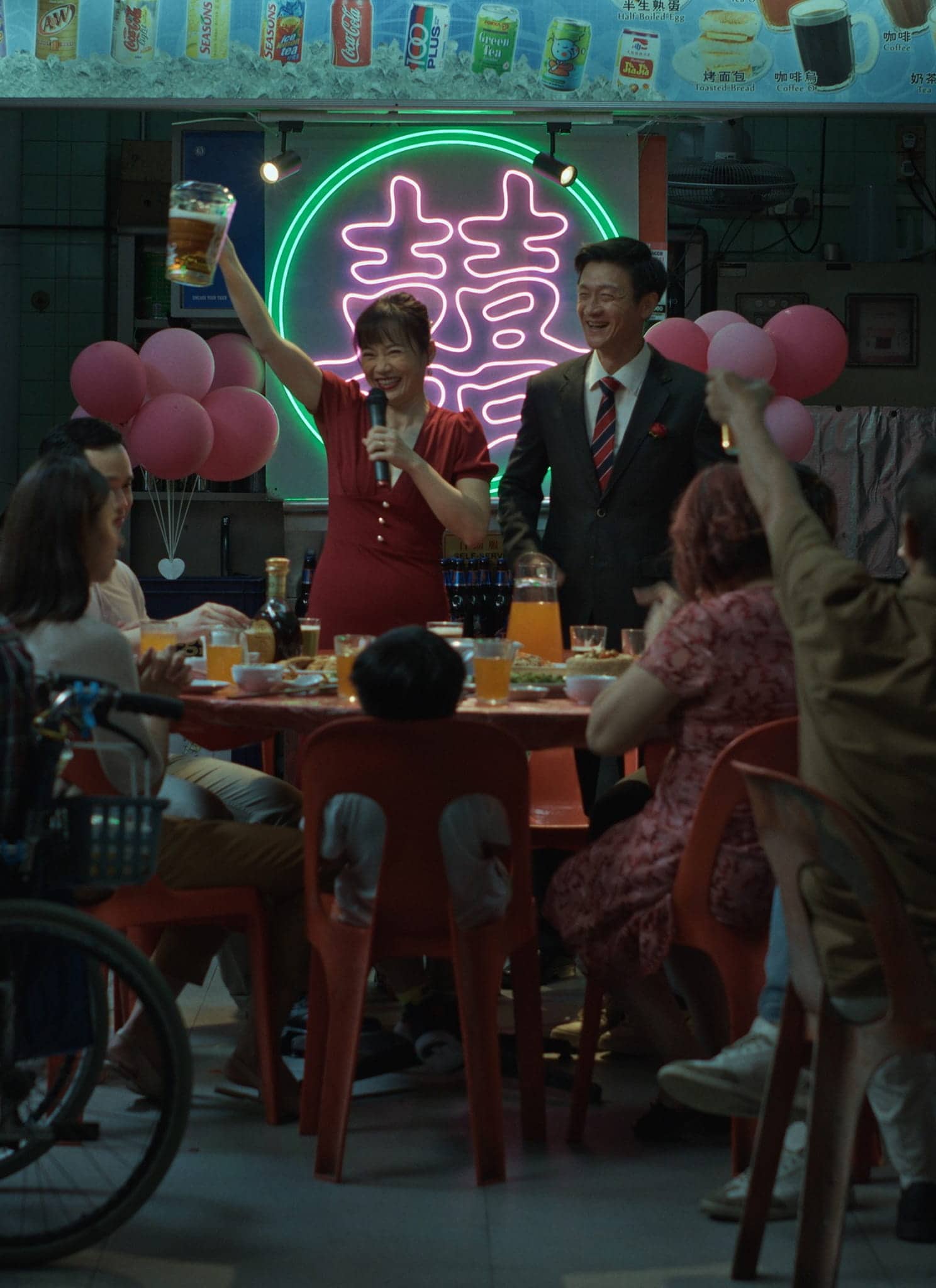 A group of people sit around tables decorated with pink balloons, raising glasses in a toast. Two people stand at the front, smiling, under a bright neon sign with double happiness symbols. The scene appears festive and celebratory.