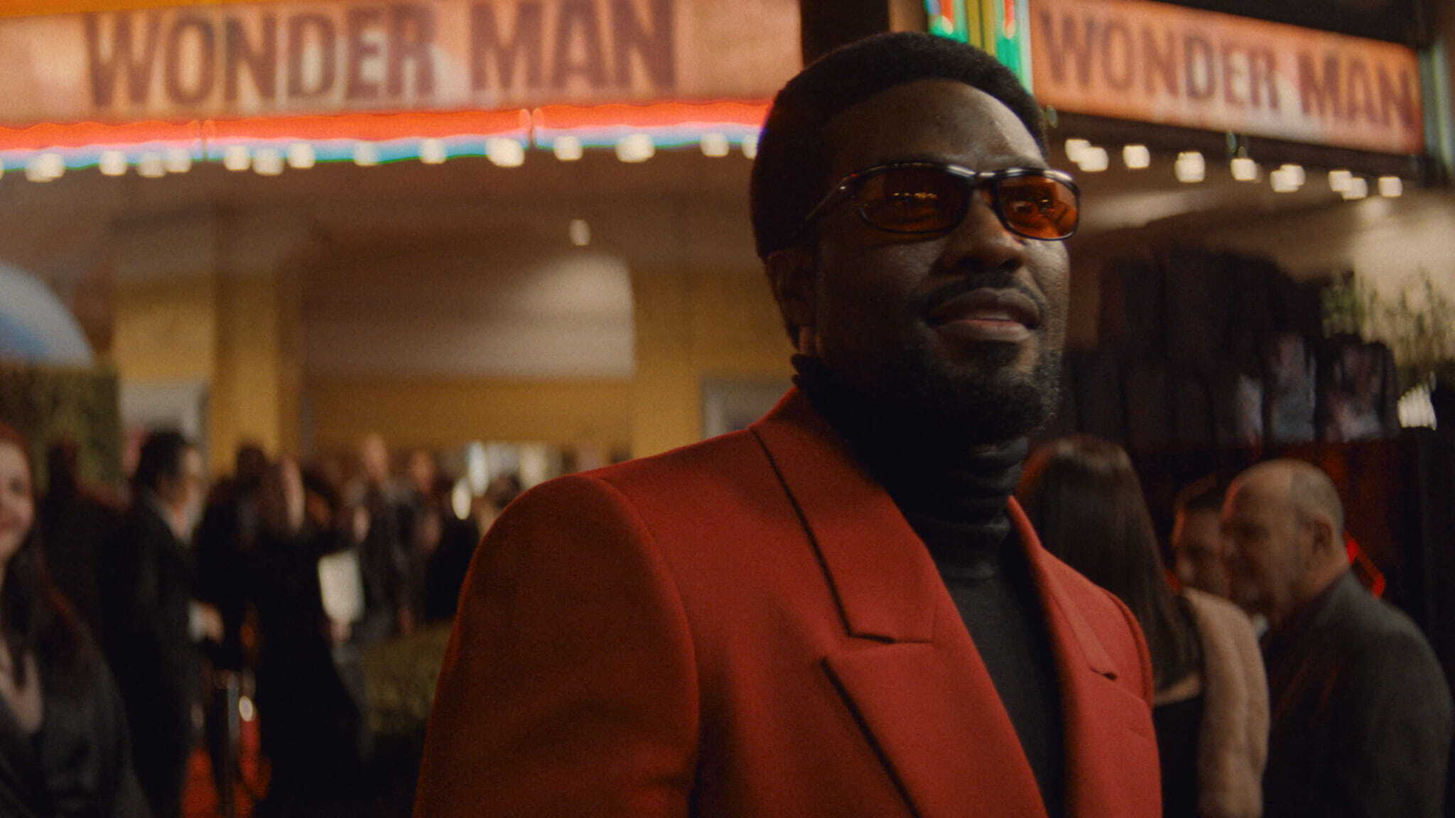 A man in a red blazer and sunglasses stands outside a theater at night. The marquee above him reads WONDER MAN, and a crowd is gathered in the background.