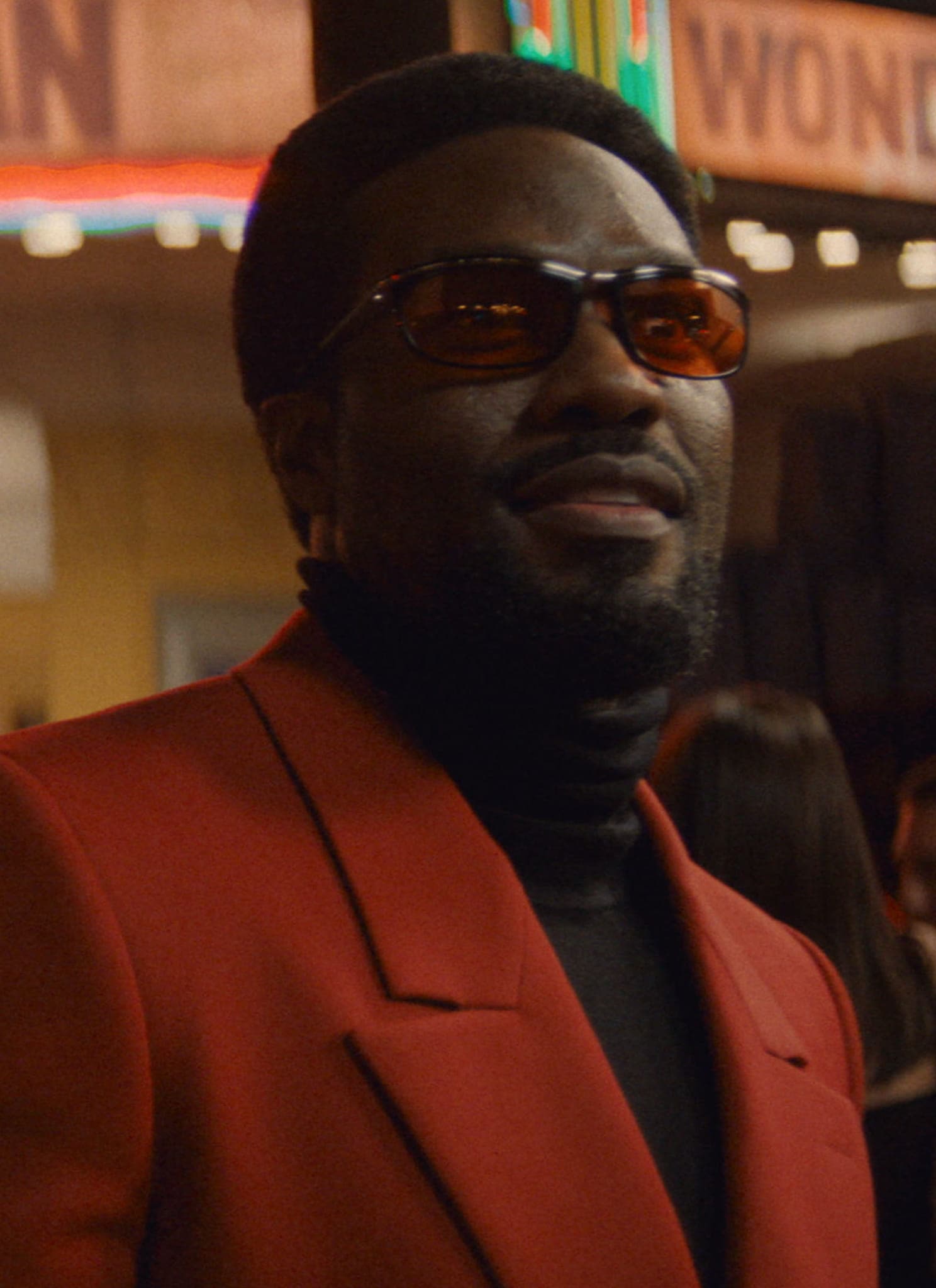 A man in a red blazer and sunglasses stands outside a theater at night. The marquee above him reads WONDER MAN, and a crowd is gathered in the background.
