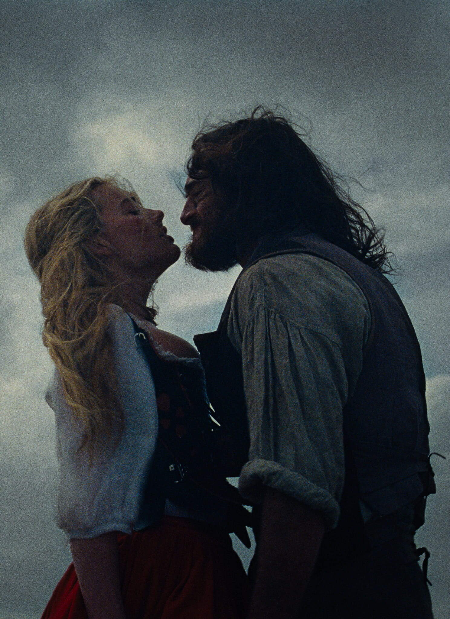 A man and a woman stand close together, gazing at each other intensely, with dark clouds looming in the sky behind them, creating a dramatic and moody atmosphere.