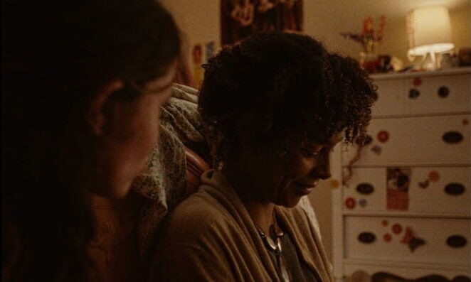 Two women sit close together in a warmly lit room. One woman, with curly hair, looks down and smiles gently; the other, partially visible, faces her. Decorated dresser and soft lighting create a cozy, intimate atmosphere.