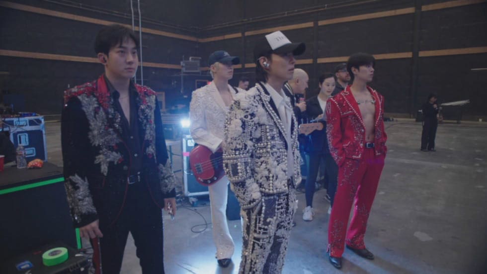 Four men from The Rose, dressed in ornate, colorful suits, stand backstage in a large, dimly lit area, appearing focused. Equipment and crew members fill the background. One holds a bass guitar while another wears a cap with ear monitors.