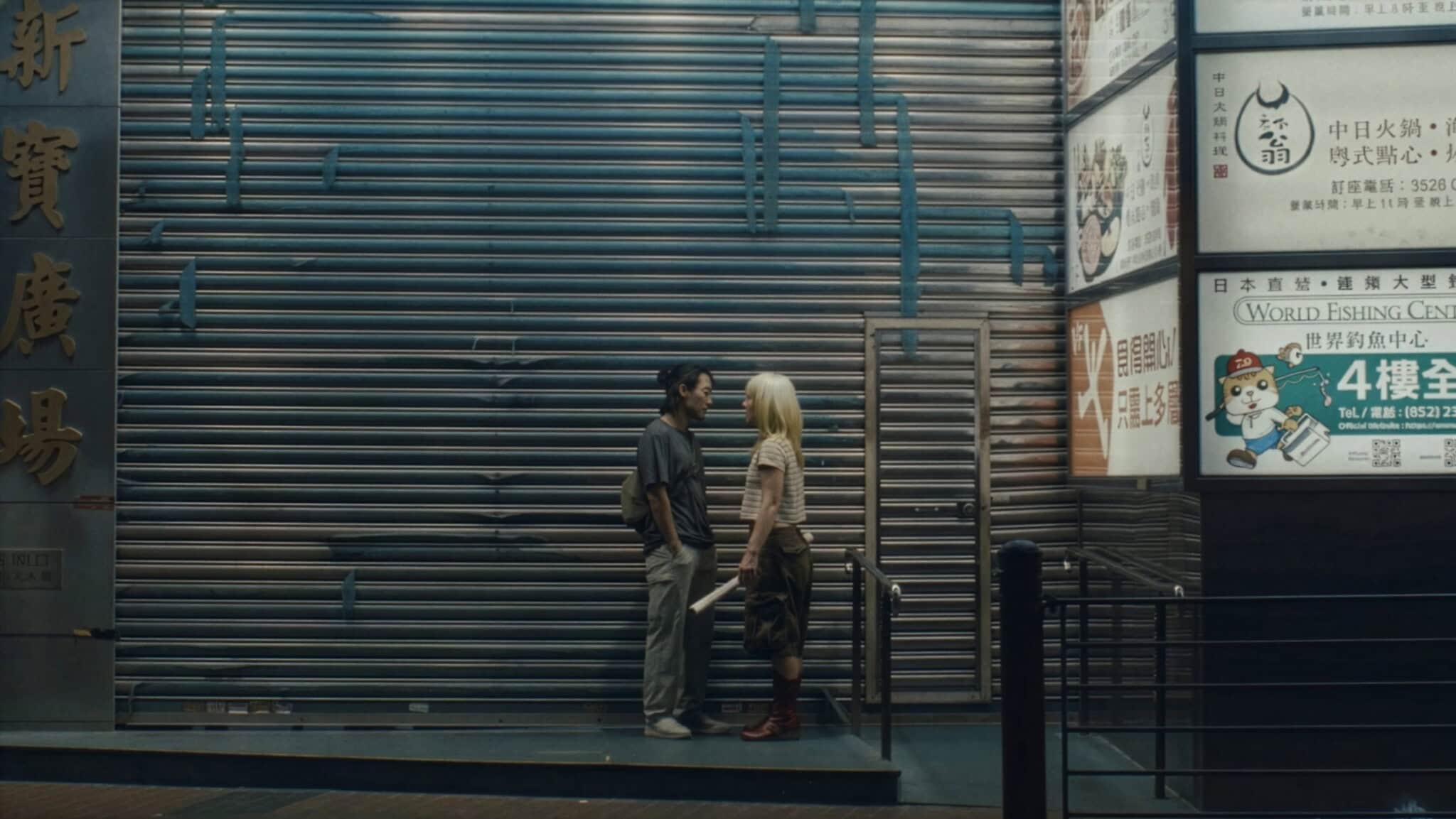 A man and a woman with platinum blonde hair stand facing each other in front of closed metal shutters at night, beneath illuminated signs with Chinese and English text in an urban setting.