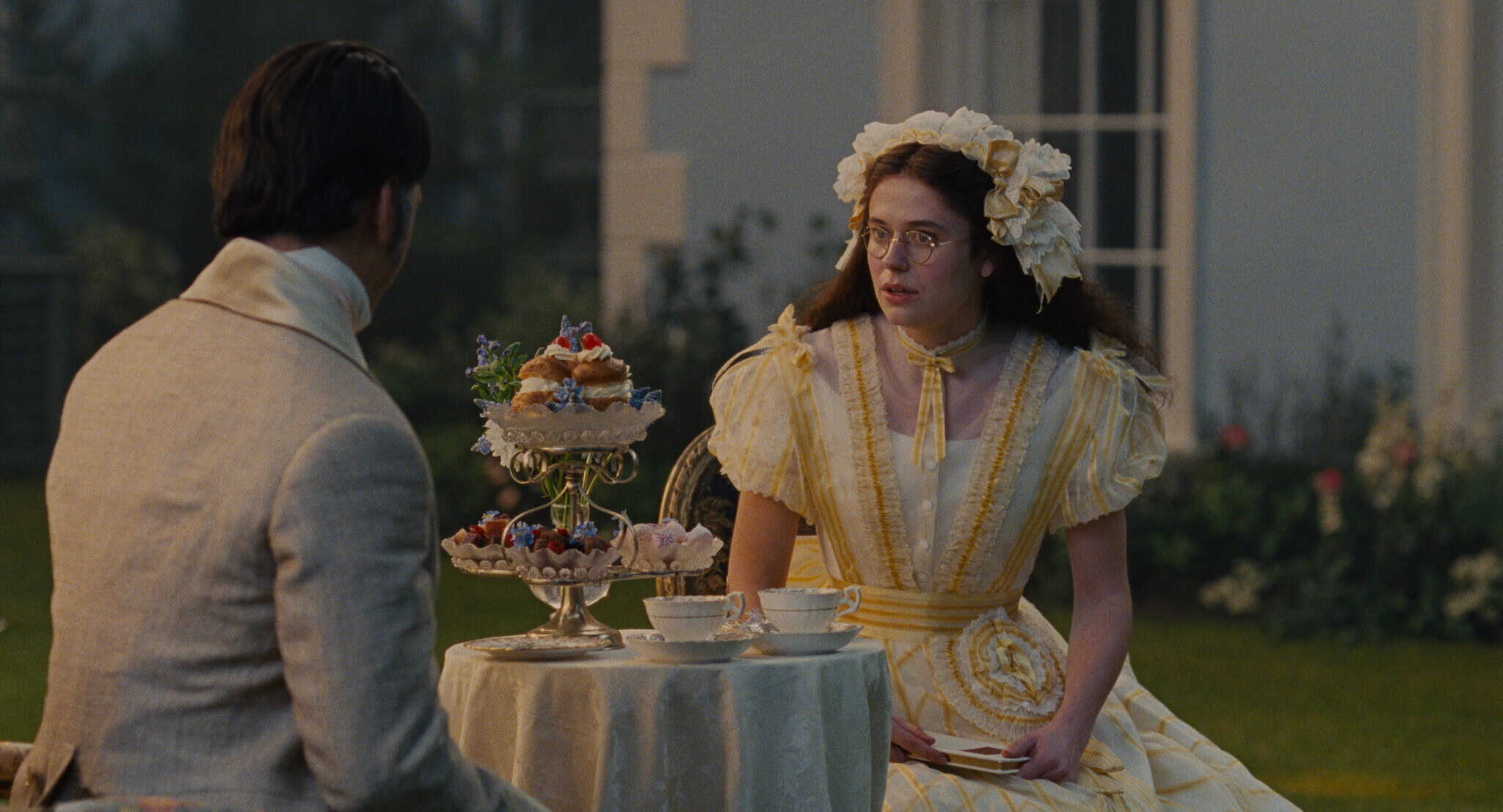 A young woman in a yellow Victorian dress and flower headpiece sits at a table with a man, facing a tiered tray of pastries and teacups, in an elegant garden setting.