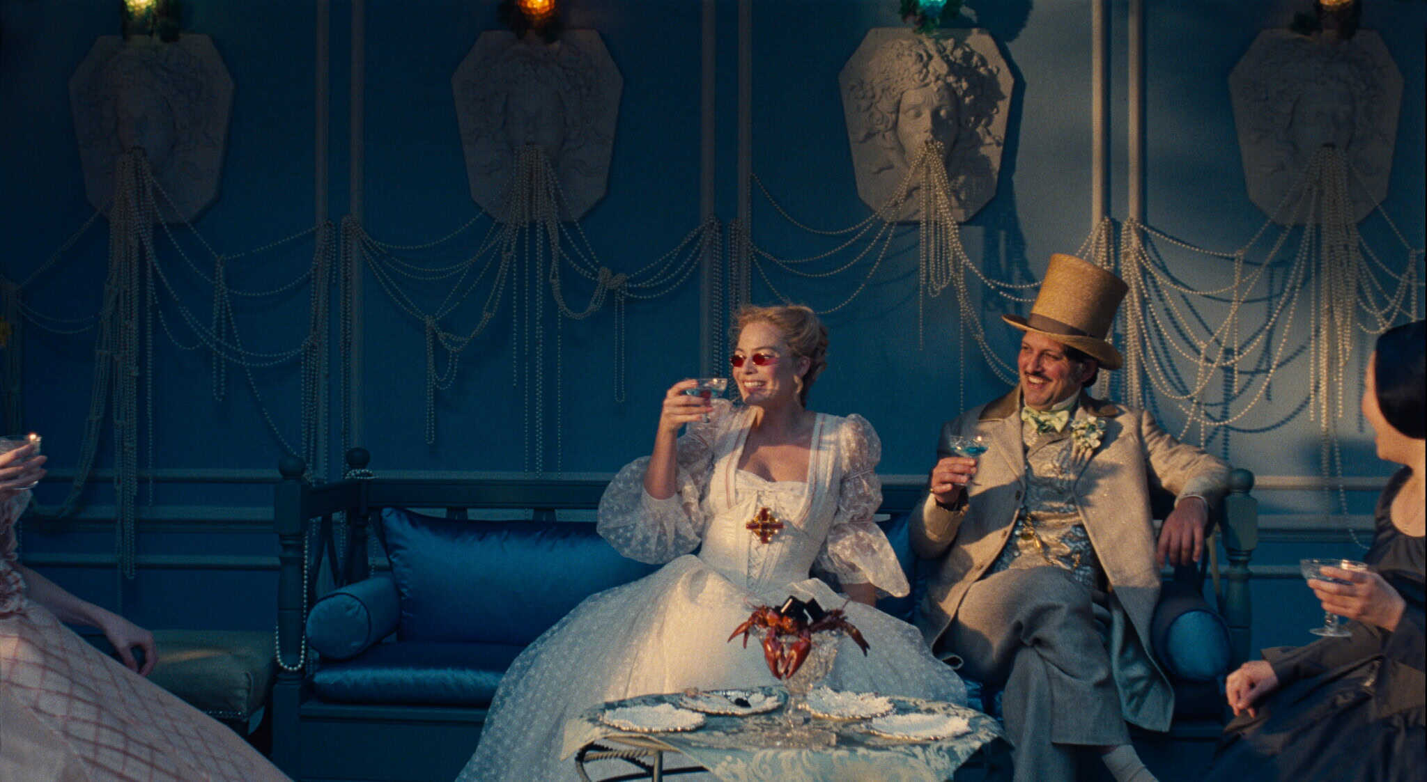 A woman in an ornate white gown and sunglasses sits on a blue sofa beside a man in a top hat and beige suit, both raising drinks, in an opulent, blue-toned room with decorative wall sculptures and string pearls.