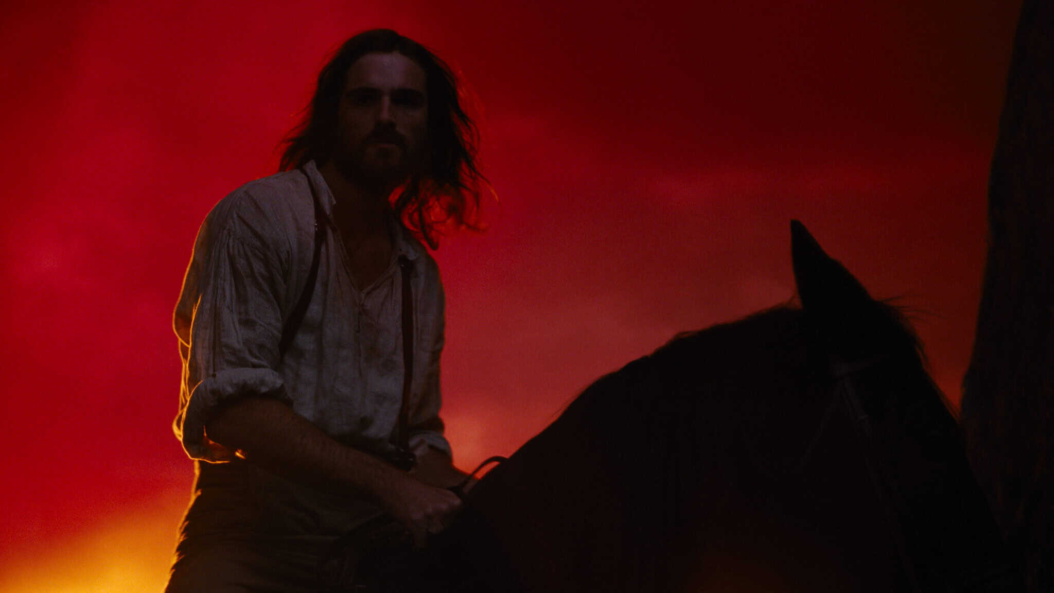 A man with long hair and a beard, wearing a white shirt with rolled-up sleeves, sits on a horse against a dramatic red and orange sky at dusk.