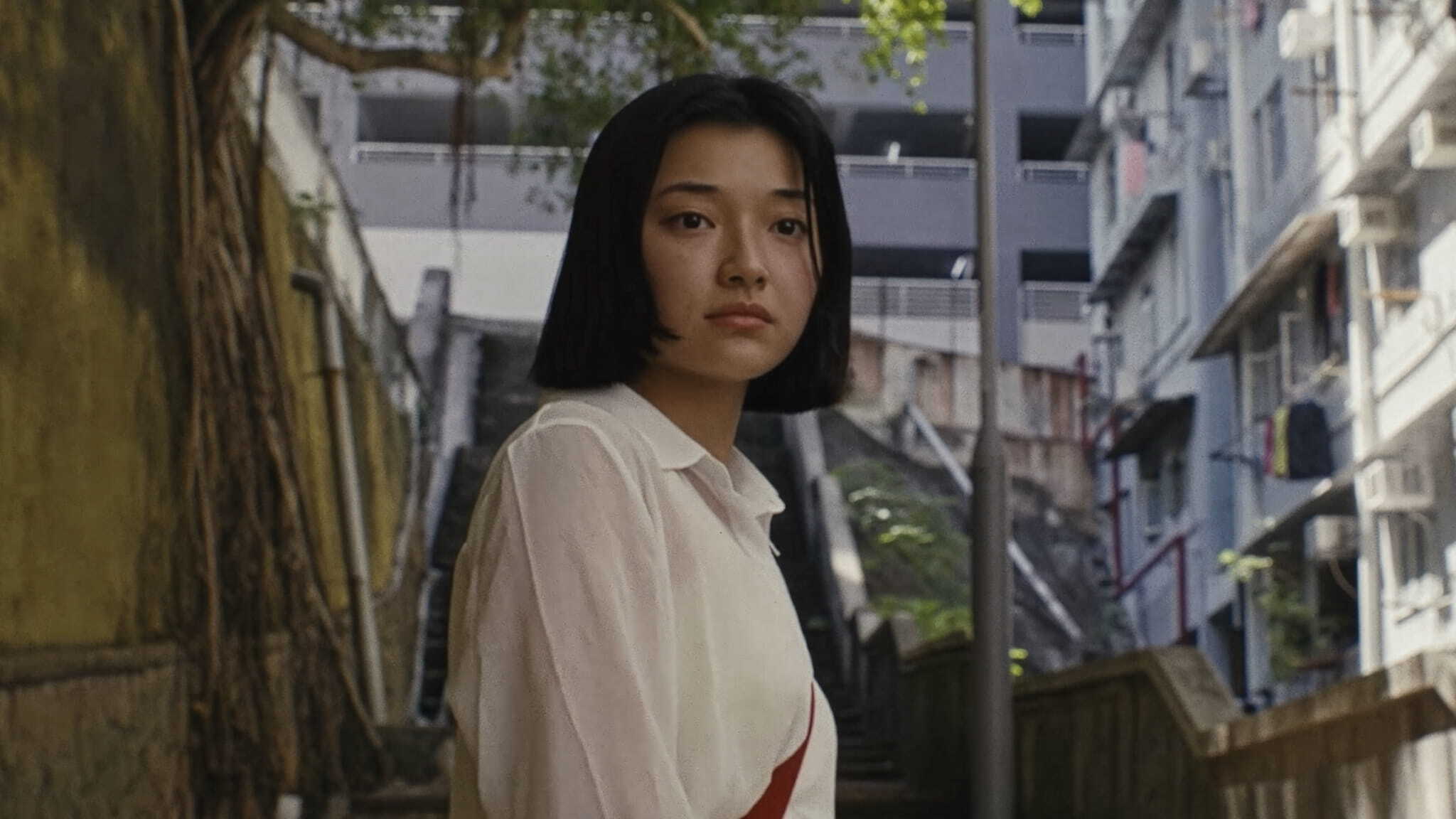 A young woman with straight black hair and a white shirt stands on an outdoor staircase between buildings, looking slightly to the side. Trees and urban structures are visible in the background.