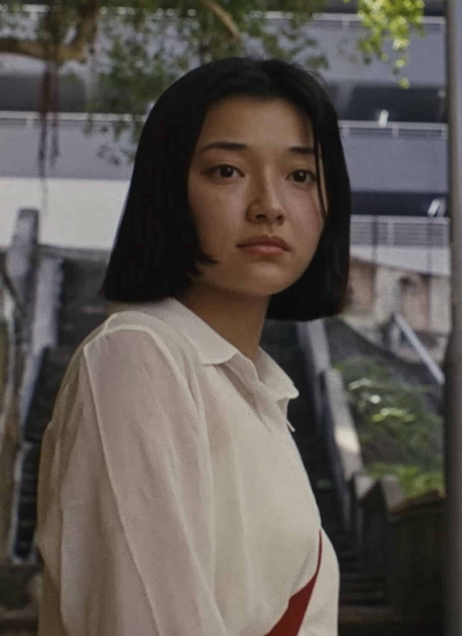A young woman with straight black hair and a white shirt stands on an outdoor staircase between buildings, looking slightly to the side. Trees and urban structures are visible in the background.