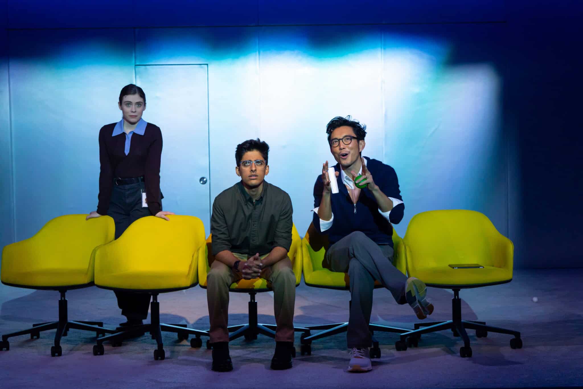 Three people on stage under blue lighting: one stands behind two others seated in bright yellow chairs. The set and background are minimalist, while the person on the right gestures animatedly, perhaps sharing insights or Karan Brar data with the audience.