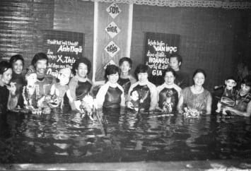 A group of people stand in waist-deep water holding traditional Vietnamese water puppets. They are smiling, and behind them are signs with Vietnamese writing on a woven wall.
