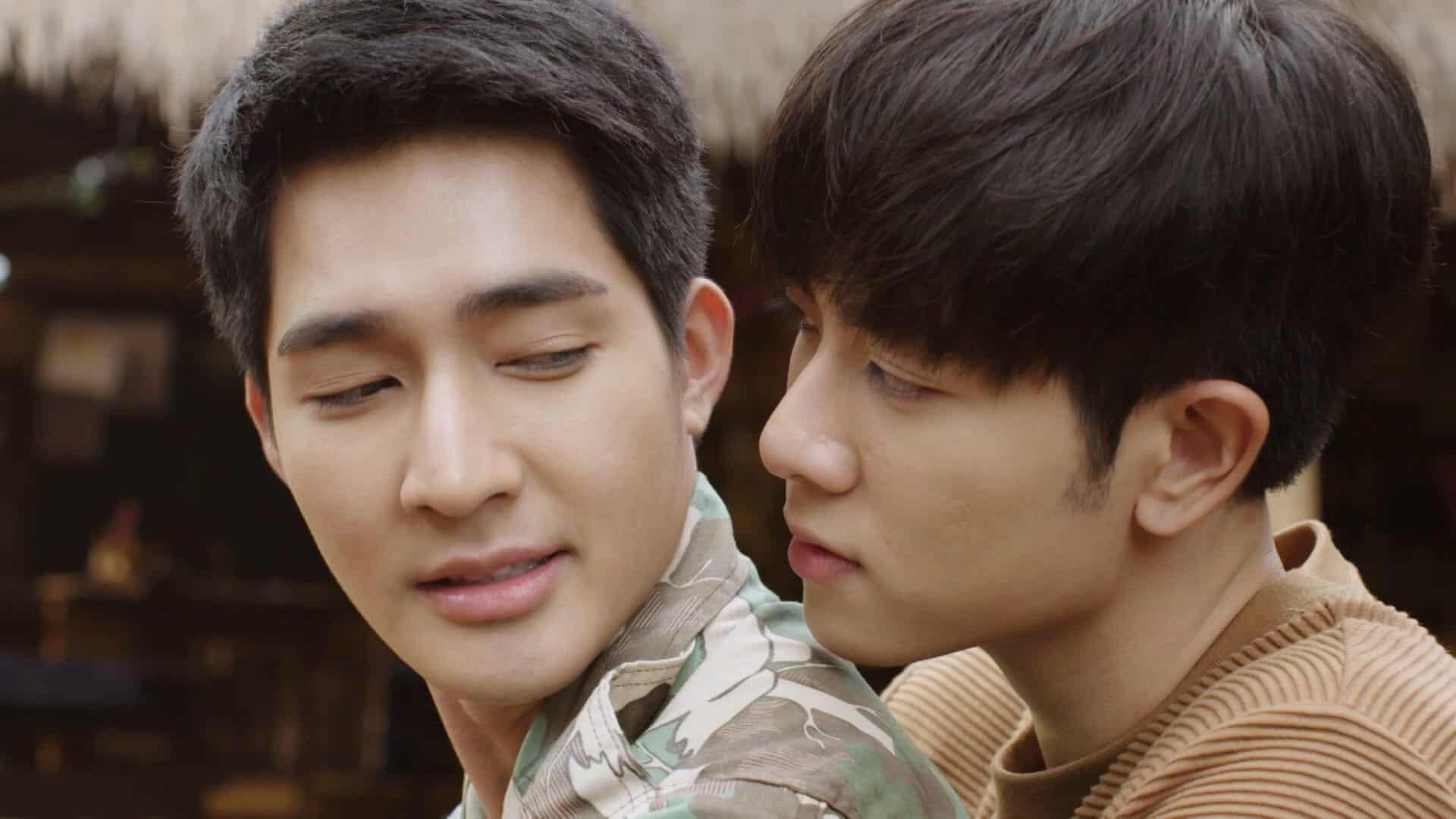 Two young men sit closely together; one in a camouflage shirt looks back with a soft smile, while the other in a brown sweater leans in, gazing at him affectionately. The background is softly blurred.