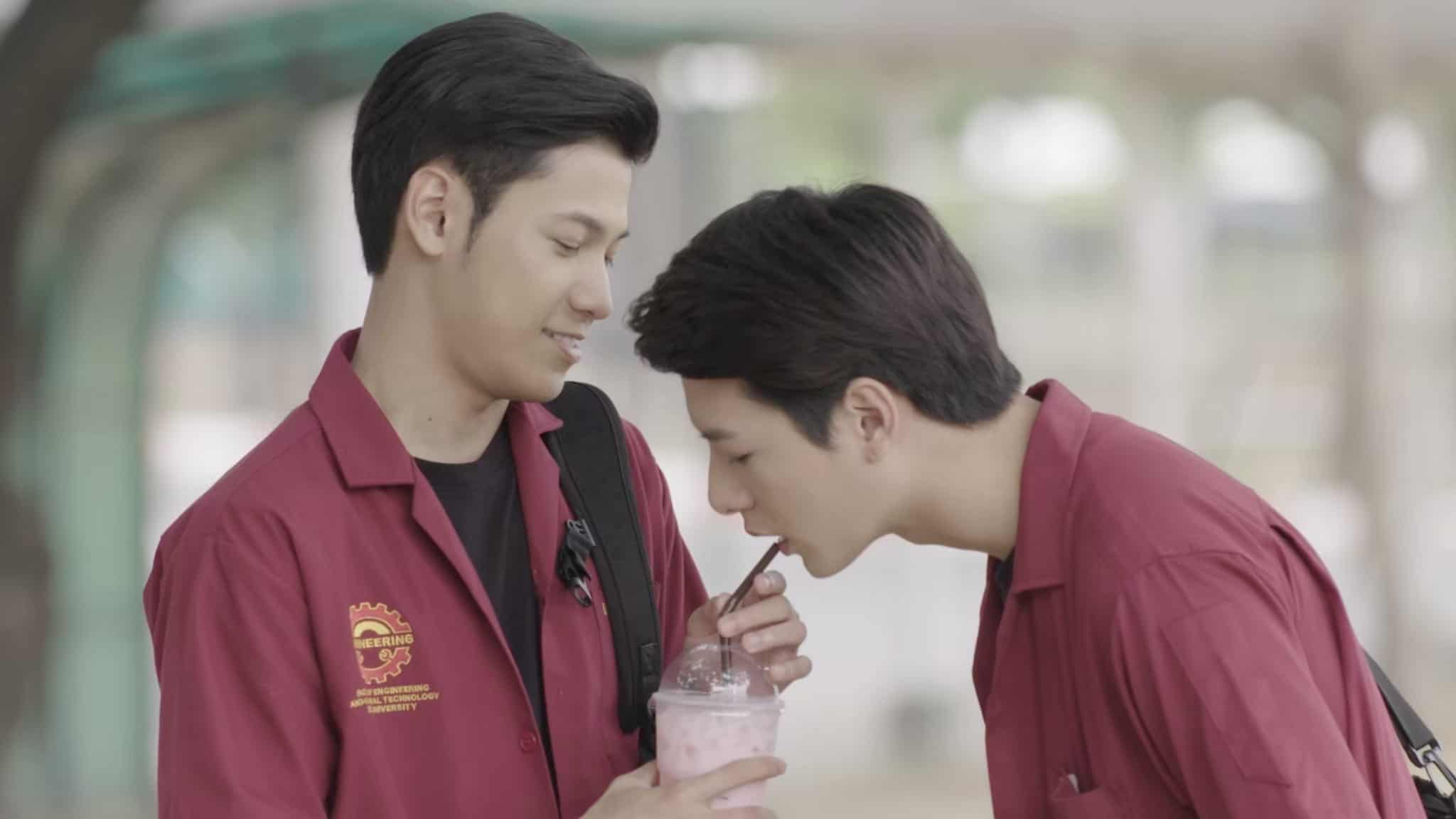 Two young men in matching red uniforms share a drink. One holds the cup and smiles while the other sips from the straw, both appearing friendly and comfortable with each other.