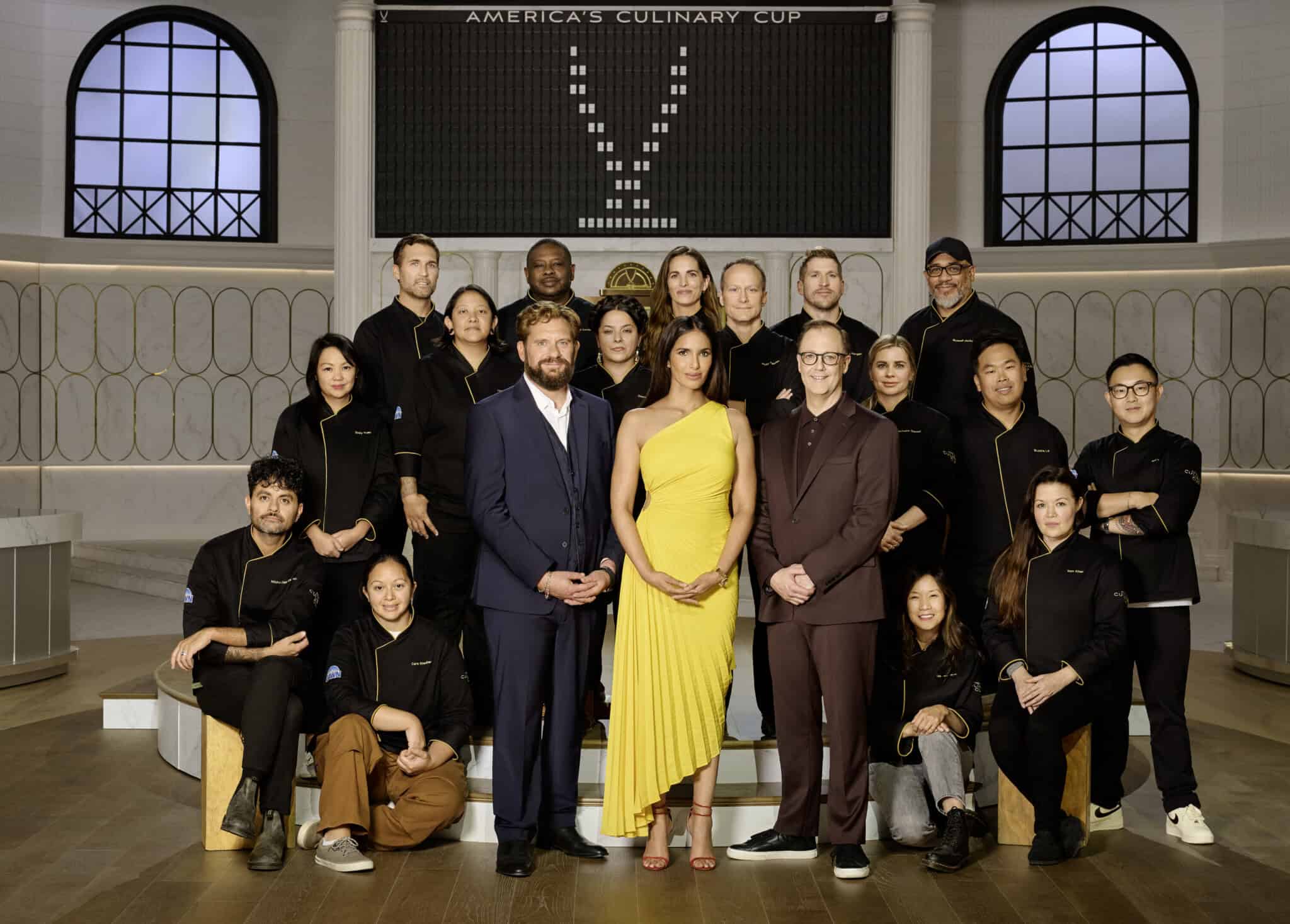 A group of people pose indoors; three people dressed formally stand at the front, while others in black chef jackets stand and kneel behind them. A scoreboard and arched windows are visible in the background.