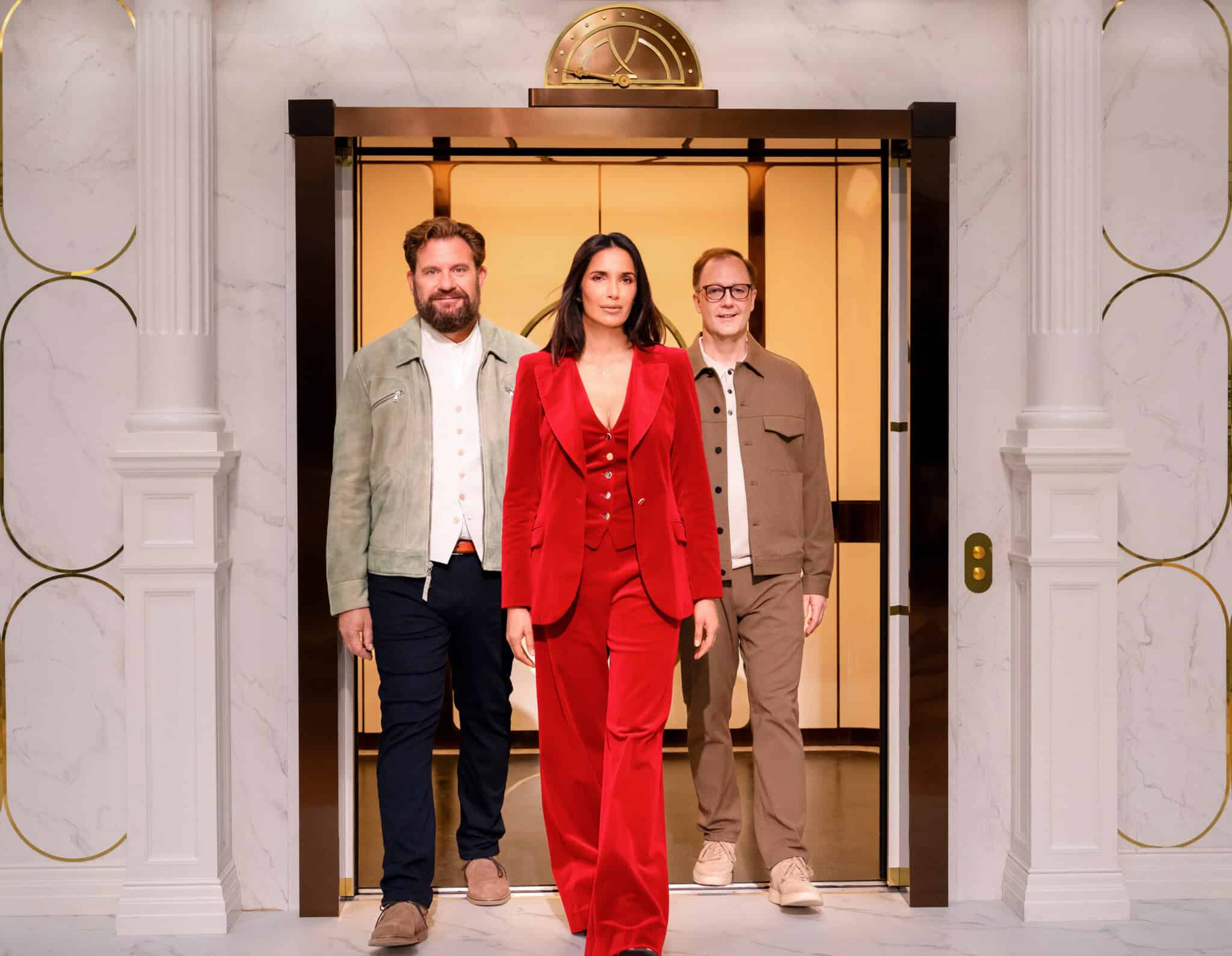 Three people stand in the doorway of a stylish elevator. The woman in front wears a red suit, flanked by two men in casual jackets and pants. The setting is elegant with marble walls and gold accents.