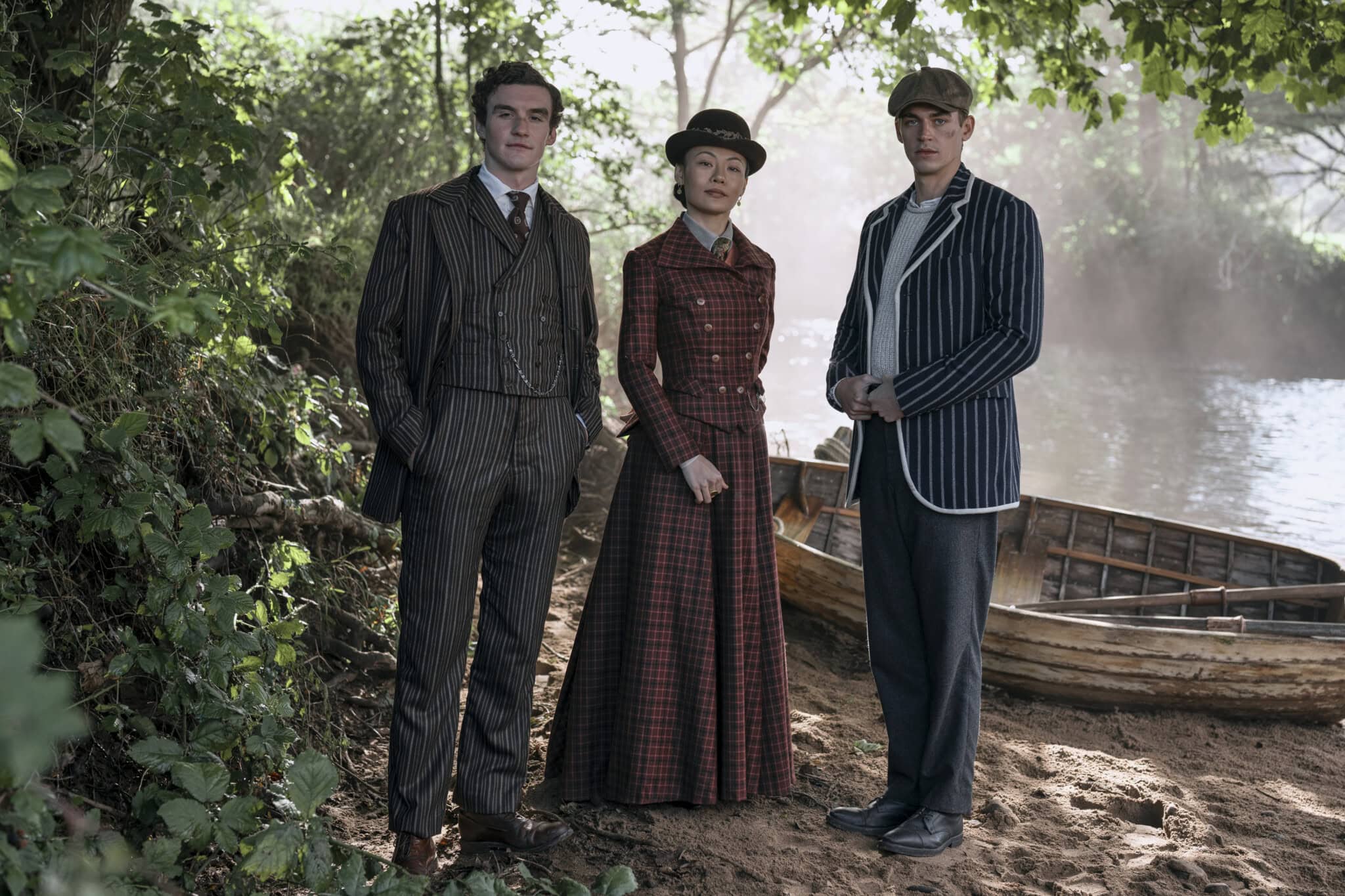 Three people in vintage clothing stand on a wooded riverbank beside a wooden rowing boat. Two men wear suits and hats, and a woman wears a long plaid dress and hat. Sunlight filters through the trees.