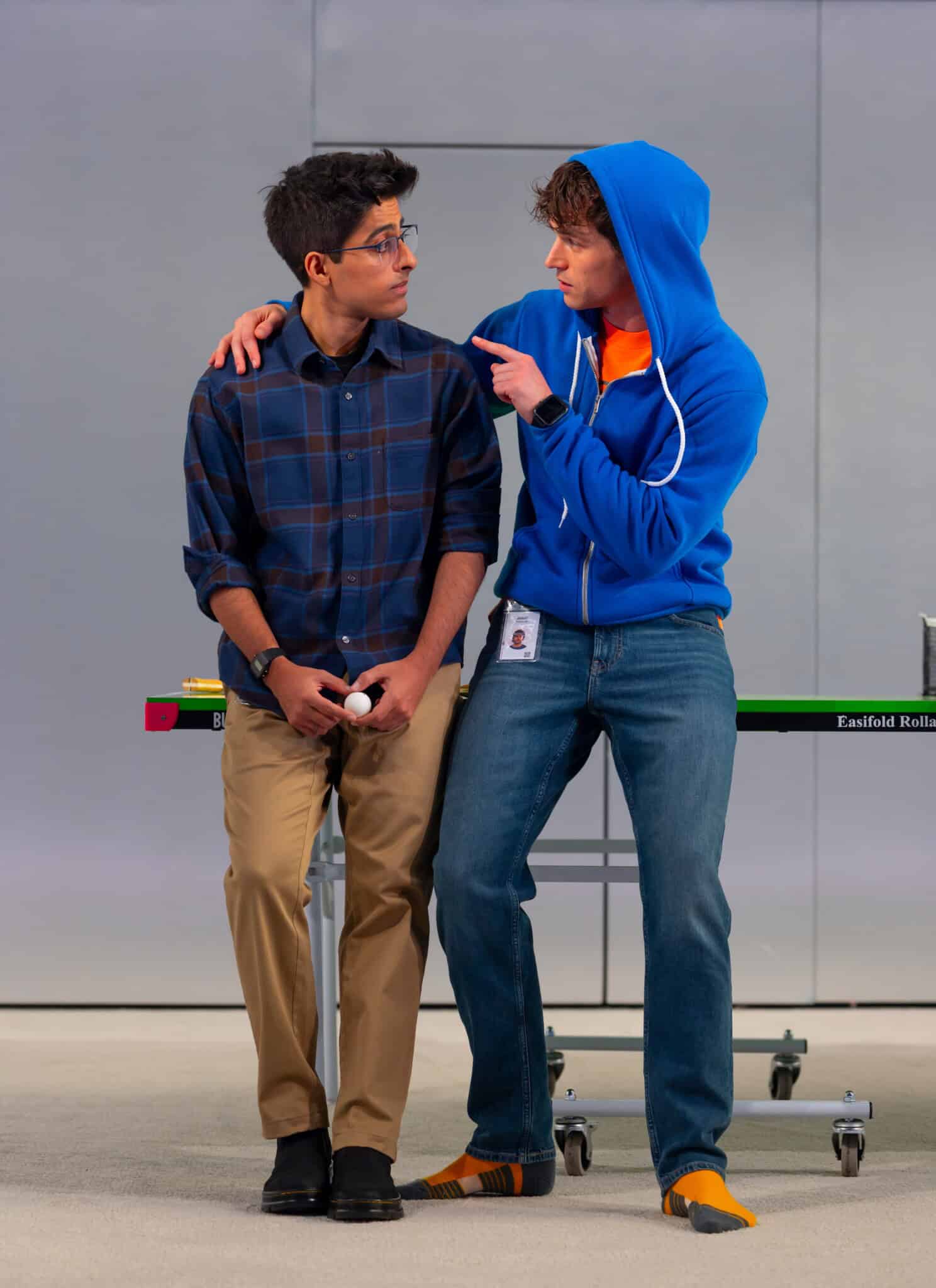 Two young men stand by a ping pong table. One, resembling Karan Brar data, wears glasses and a plaid shirt holding a ball; the other, in a blue hoodie and jeans, has his arm around him and is pointing while talking to him.
